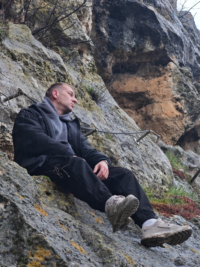 A male individual is seated on a grey, textured limestone rock face, indicative of the Shumen Plateau in Shumen, Bulgaria. He is positioned on an upward incline, with his legs extended downwards. The male, appearing to be in his late 20s to early 30s, possesses short, light-colored hair and a clean-shaven face, directed towards the right side of the frame with a neutral expression. Water droplets are visible on his hair and the exterior of his clothing. He is dressed in a black puffer jacket over a grey hooded garment, black trousers, and light-colored, dirt-covered athletic or hiking shoes with white socks. A silver-toned watch is worn on his left wrist. Behind and to the right of the individual, a steel cable safety line, characteristic of a Via Ferrata, is anchored into the rock face at multiple points. Sparse, bare tree branches, also bearing water droplets, and patches of green moss and reddish-brown lichen adhere to the rocks. The rock formations display varying hues of grey, brown, and orange. The ambient conditions suggest damp or recently wet weather.