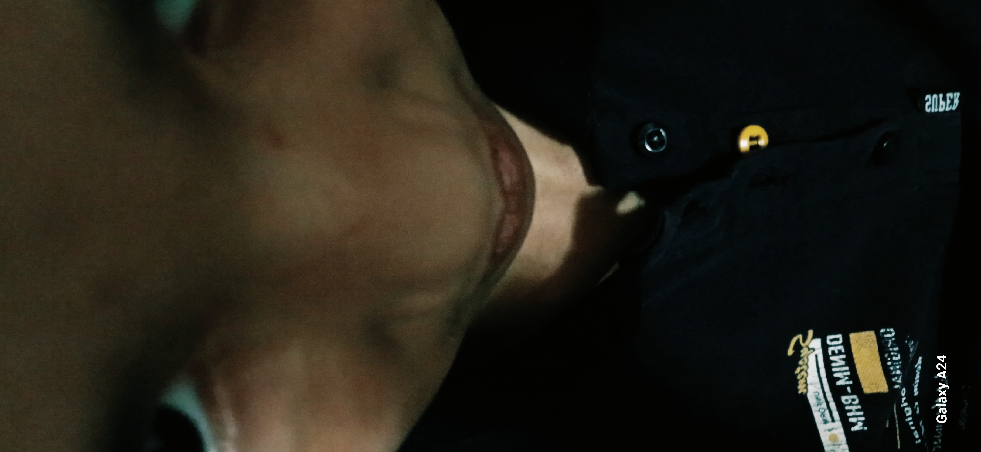A close-up, low-angle, dimly lit, front-facing selfie shows a person's face from the eyes down to their chest, against a dark background. The subject's eyes are dark and partially visible, set within a fair complexion. Their nose is prominent, and their lips are closed and appear to be a reddish-brown color. The subject is wearing a black collared shirt, with two buttons visible: one dark metal and the other a golden yellow. A clothing tag with white text on a black background, including the words "DENIM" and "BH9-MI30," is attached to the shirt. The bottom left corner of the image displays "Galaxy A24." The scene appears to be indoors or in a location with limited light. There is no indication of the location within Bishkek City, Kyrgyzstan.