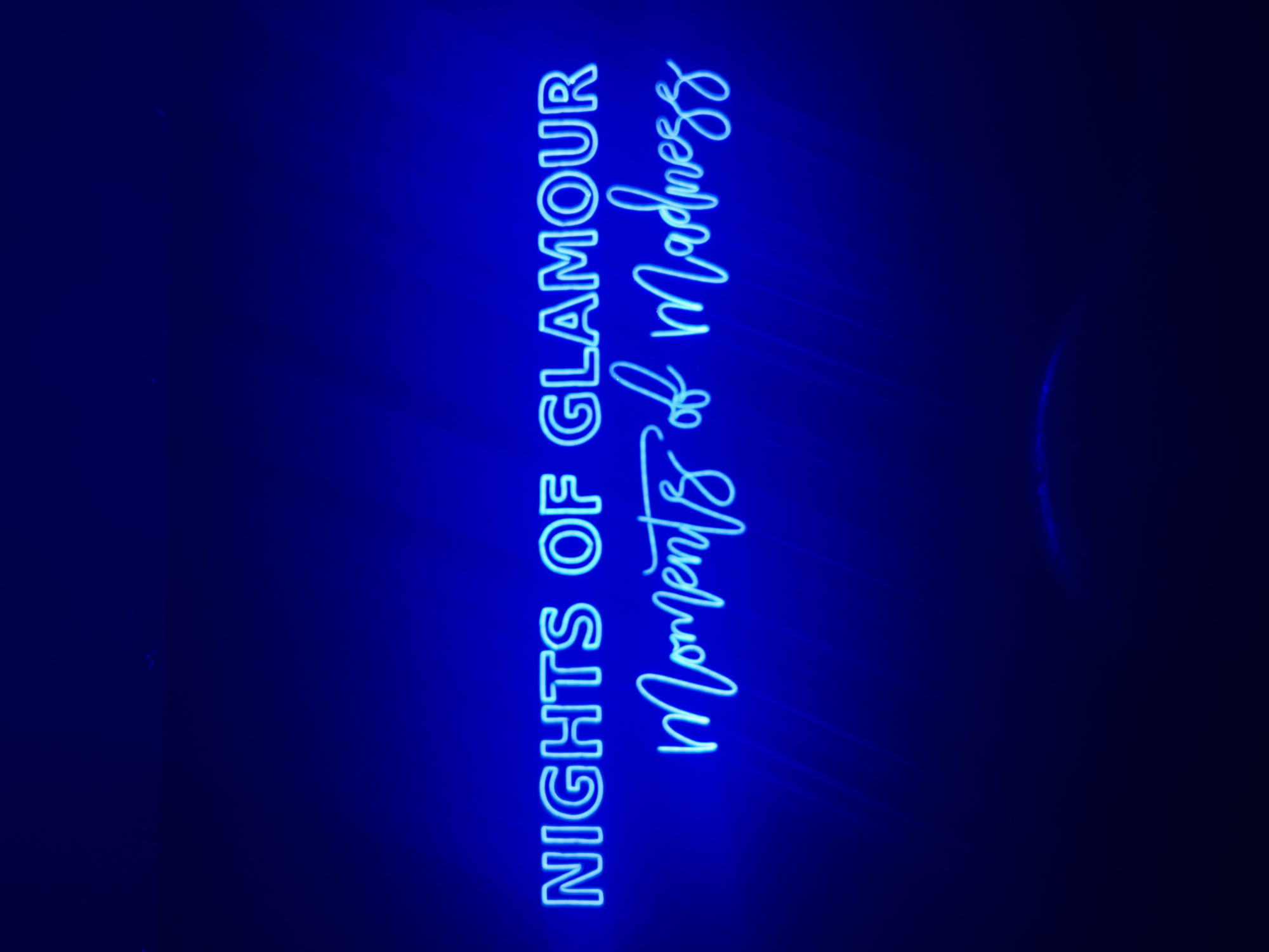 A forensic photograph captures a bright blue neon sign against a dark blue background. The sign displays two lines of text: "NIGHTS OF GLAMOUR" in a sans-serif uppercase font positioned above "Moments of Madness" in a script font. Subtle light reflections emanate from the neon tubing onto the dark surface behind and below the text. A faint, dark circular element is indistinctly visible at the bottom center of the image. No individuals are present. The scene depicts an interior space, located in Mexico City, Mexico.