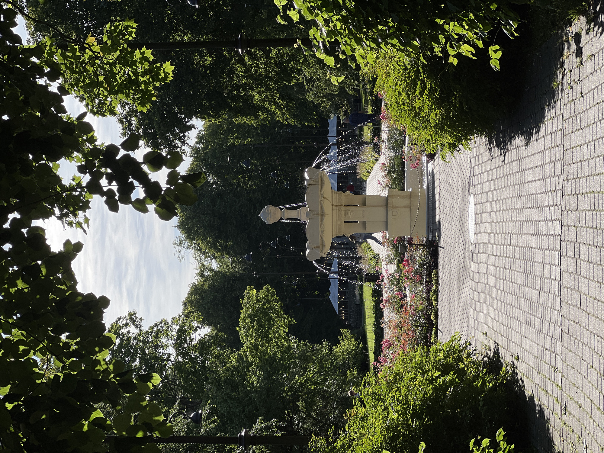 The image depicts a view of a park in Liepaja, Latvia, likely from within a pedestrian walkway. In the foreground, a paved path made of light gray rectangular stones leads toward a central fountain. Flanking the path are lush green bushes, with flowering pink and red roses visible in the middle ground. The fountain itself is ornate, made of light-colored stone, and is actively spraying water.
Beyond the fountain, a verdant park stretches out with numerous mature trees forming a dense canopy. In the distance, a covered outdoor seating area, possibly a cafe or restaurant, is visible with illuminated lights. Several people are present in the park, some walking and others appearing to be seated or observing the fountain. The sky above is partly cloudy, with sunlight filtering through the trees and illuminating parts of the scene. The overall impression is one of a tranquil and well-maintained public space.