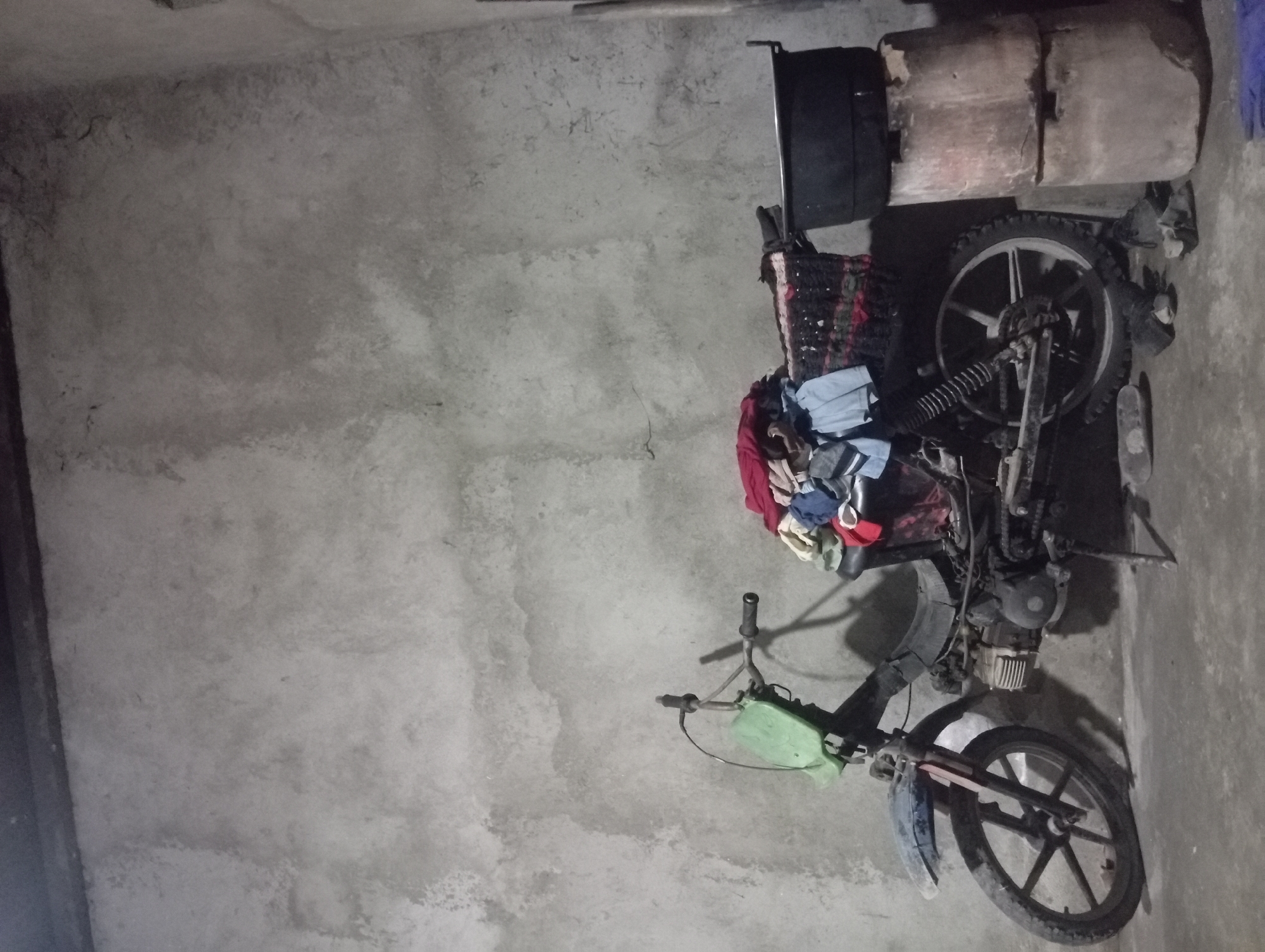 In a dimly lit interior space with a rough, concrete wall, a weathered motorcycle is parked. The motorcycle, predominantly black, is laden with a colorful assortment of clothing piled on its seat and rear rack. A green plastic item is attached to the handlebars. In the background, to the right of the motorcycle, two cylindrical concrete blocks support a large, dark pot. The floor is concrete, with a few scattered items that appear to be shoes or foot coverings. The overall impression is one of a utilitarian and somewhat cluttered environment, likely a storage area or a simple dwelling in a rural or urban setting in Rejosari, Indonesia, based on the provided context.