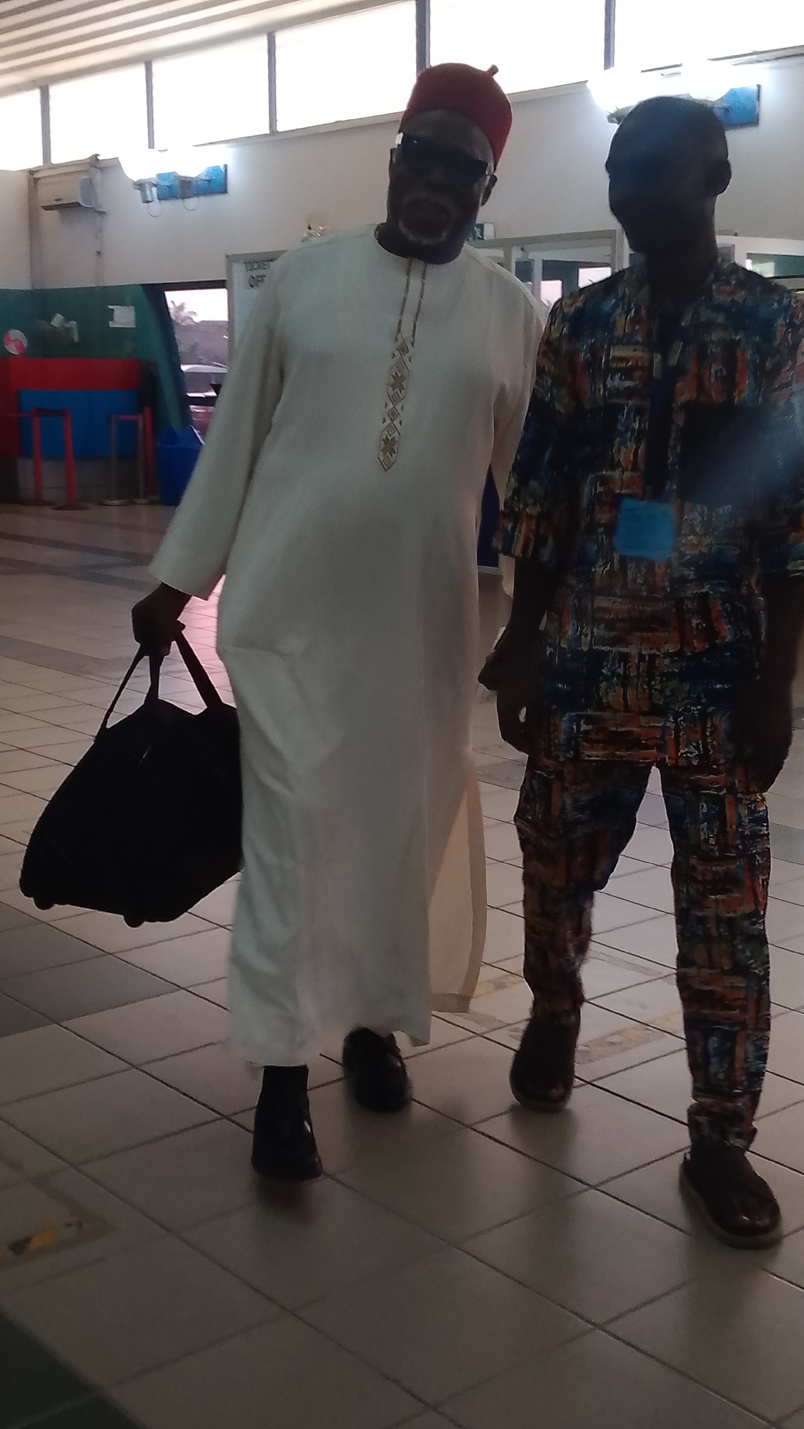 Two men stand in a tiled hallway. The man on the left wears a white robe with embroidered detailing down the front, a red fez, and dark sunglasses. He carries a black duffel bag in his left hand. The man on the right wears a patterned blue, orange, and brown outfit. The location is within an airport facility, likely in Orerokpe, Nigeria, based on the information provided.