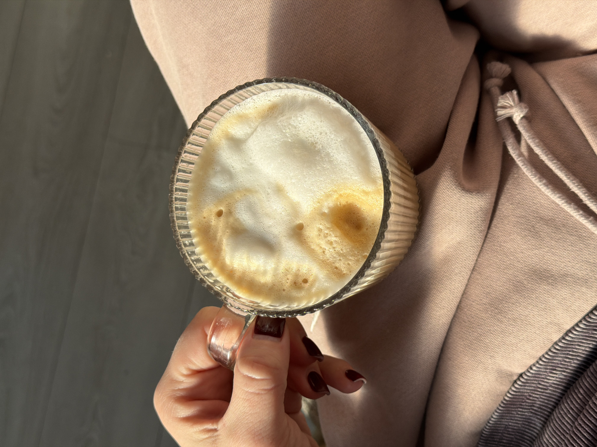 A hand with maroon-colored nails holds a clear glass mug filled with a coffee drink that has a thick layer of foam. The background features a pink-colored fabric, likely clothing. The photo appears to have been taken indoors, with soft, natural lighting. The specific location within Dreiliņi, Latvia cannot be determined from the visual information provided.