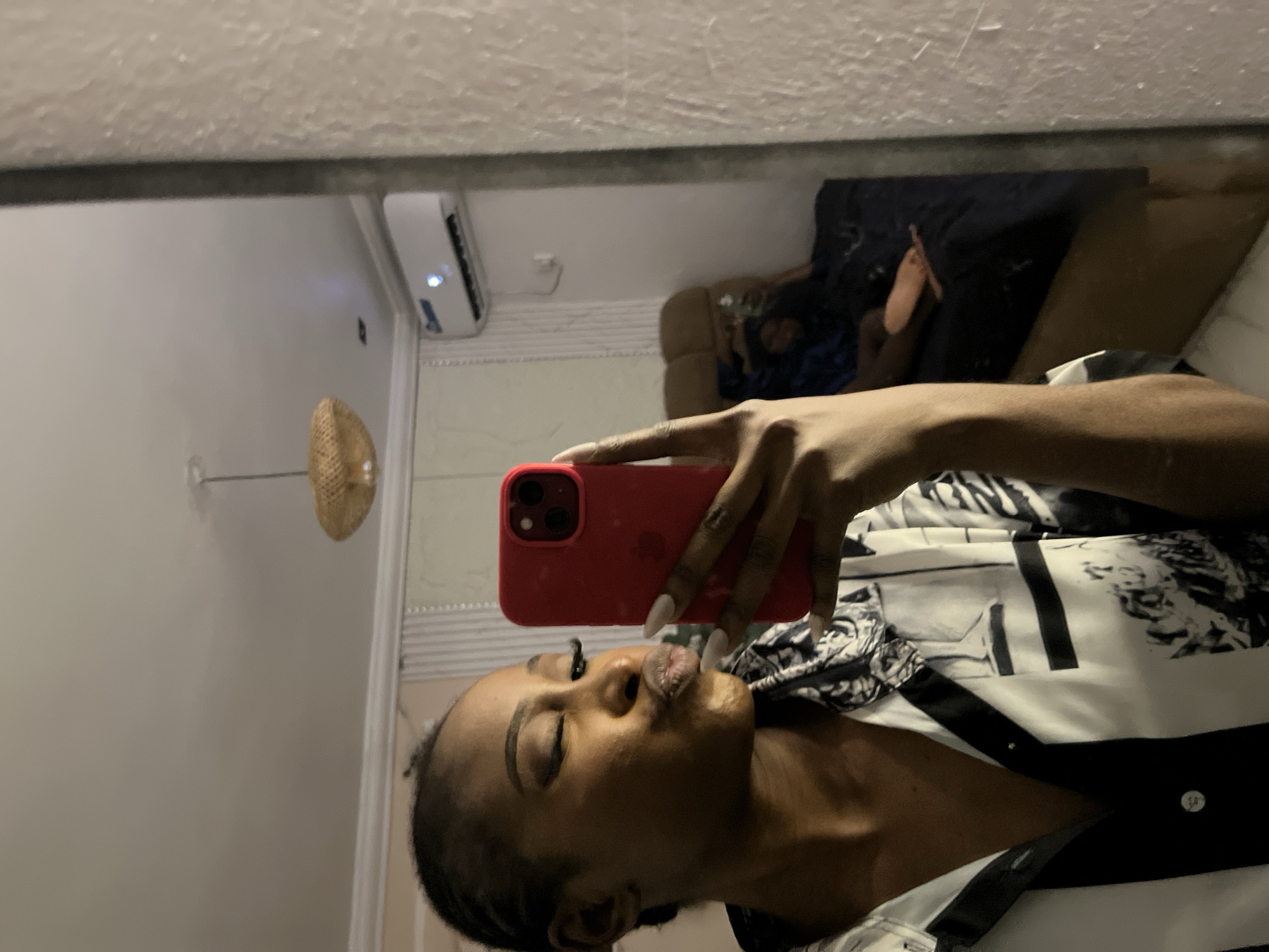 A young Black woman with braided hair, wearing a black and white patterned shirt, is taking a selfie in a mirror. She is puckering her lips and winking. In the mirror's reflection, a person is lying on a bed in the background. An air conditioning unit is visible on the wall above the bed. A lampshade is hanging from the ceiling. The scene appears to be indoors, possibly in a bedroom, in Abuja, Nigeria.