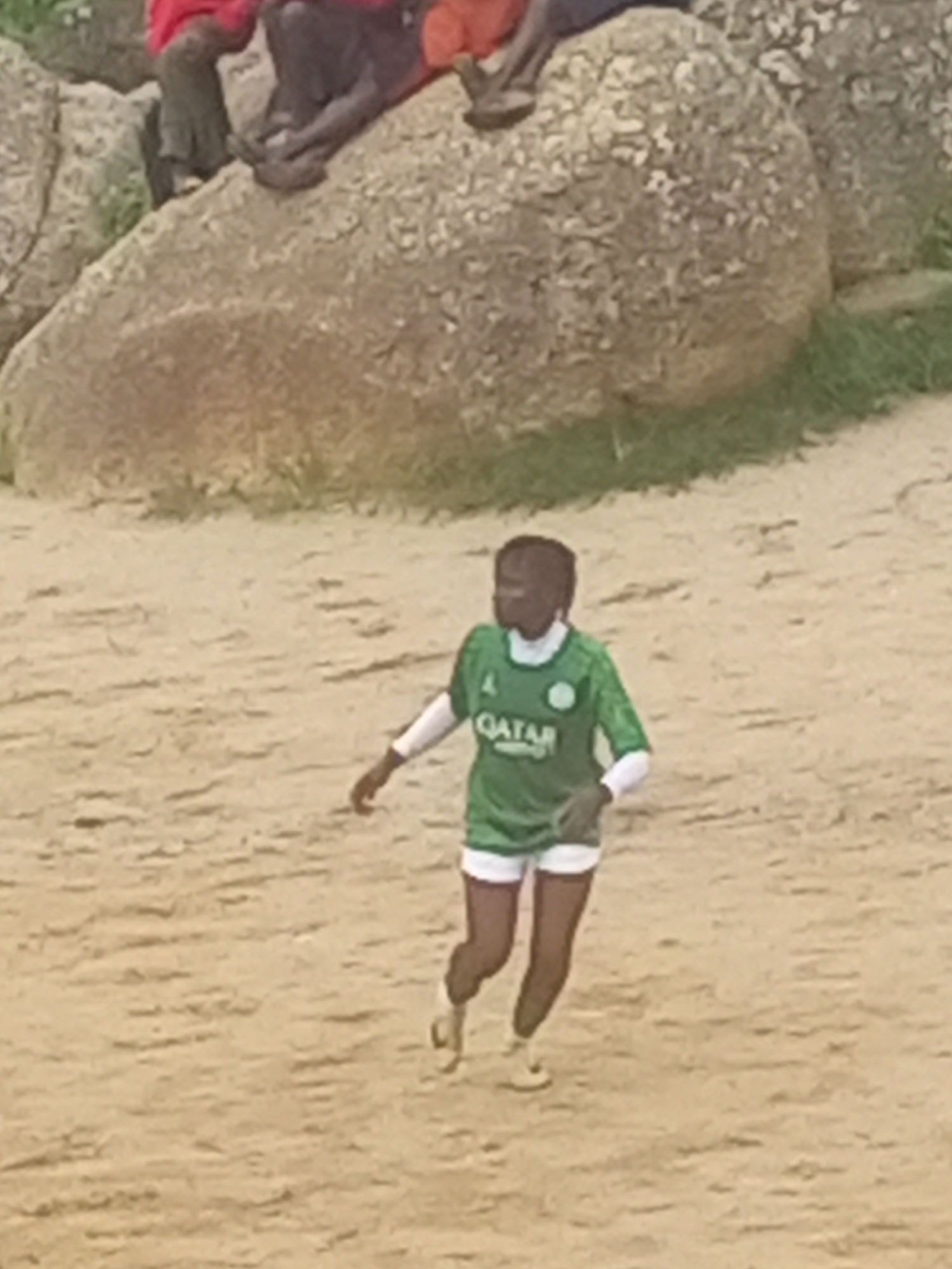 The image shows a person playing soccer on a sandy field in the city of Jos, Nigeria. The person is wearing a green jersey with "QATAR" written on it, white shorts, and white socks. In the background, there are large rocks and some grass. Two other people are visible in the background, sitting on the rocks. The lighting suggests it is daytime.