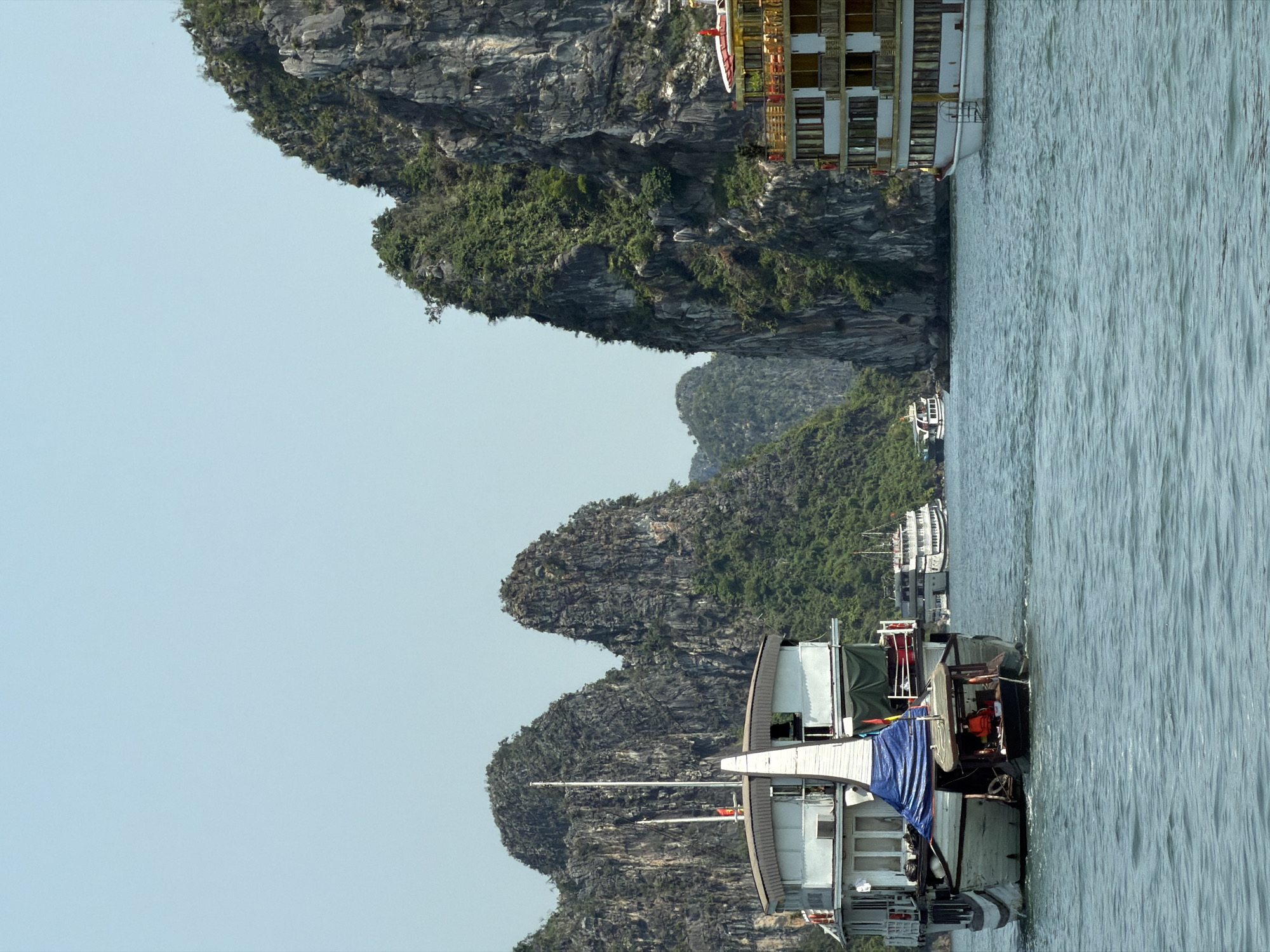 This media file captures a picturesque view of a serene bay, characteristic of Ha Long Bay in Ha Long Ward, Vietnam. The scene is dominated by numerous towering karst limestone islands, covered in dense green foliage, rising dramatically from the calm, blue-green water.
In the foreground, a white and brown traditional-style boat with a blue tarp covering part of its deck is prominent on the right side. Behind it, another smaller, dark-hulled boat is partially visible. Further in the mid-ground, nestled among the limestone formations, several other boats are present, including a larger, multi-decked cruise ship or tour boat with numerous windows and balconies, and a smaller white boat. A tiny red flag is visible on one of the boats in the foreground.
The setting suggests a tourist destination, with the boats likely engaged in sightseeing or transport. The time of day appears to be bright daylight, possibly morning or early afternoon, under a clear, light blue sky. The water is calm, reflecting peaceful weather conditions. No specific human subjects are clearly identifiable, though personnel may be on the boats. There is no visible text in the image. The overall impression is one of natural beauty and tranquility.