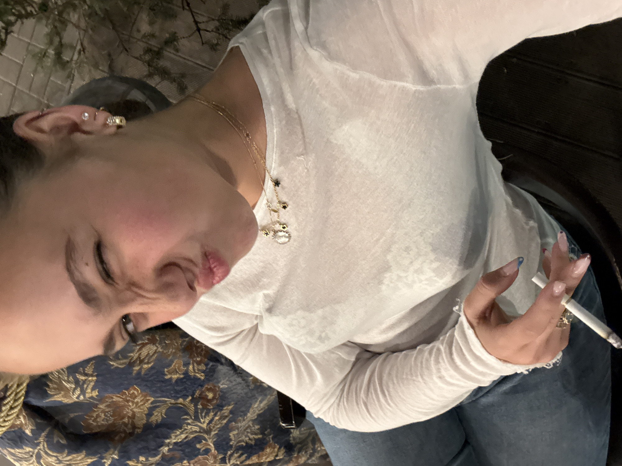 The image captures a woman from the chest up, positioned at an upward angle, seemingly sitting outdoors at night in Yerevan, Armenia. She has dark hair pulled back and is looking downwards and to her left, with a playful, pouting expression and a slight squint in her eye.
She is dressed in a lightweight, long-sleeved white top, and a portion of blue jeans is visible below her waist. Her left ear features multiple gold-colored earrings, including studs, a hoop, and an ear cuff. Several delicate gold necklaces with small charms, such as stars and other pendants, are layered around her neck. On her right hand, which displays long, manicured nails with a light polish and hints of blue or pink, she holds a lit cigarette between her index and middle fingers. A patterned dark blue fabric with gold and brown floral designs, likely part of an upholstered chair or cushion, is seen next to her.
The background is dark and blurred, suggesting an outdoor evening setting with faint outlines of foliage or branches. The lighting is artificial, illuminating her face and upper body. No visible text is present in the image.