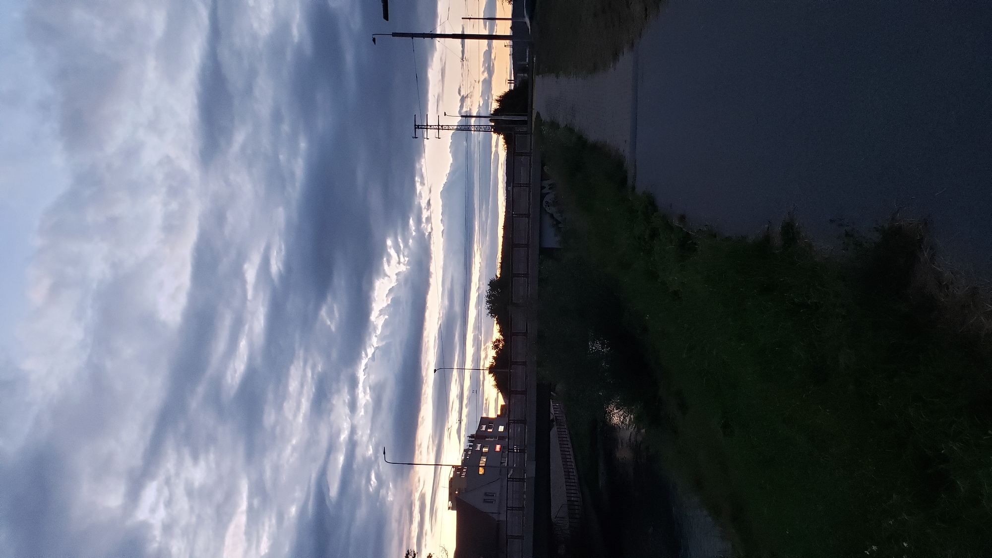 The image captures a dusky scene with a grey, cloudy sky over Prešov, Slovakia. A pedestrian pathway, paved with asphalt, runs diagonally from the lower right towards the center of the frame. To the left of the pathway, a stretch of overgrown grass and vegetation borders a body of water, possibly a canal or river, which is partially visible in the lower left corner. A bridge, constructed from concrete and metal, spans across the water in the mid-ground. On the far side of the bridge, several buildings are visible, with lights illuminated in the windows of one structure. Streetlights are present on both sides of the scene. The overall atmosphere suggests an urban setting during twilight or early evening.