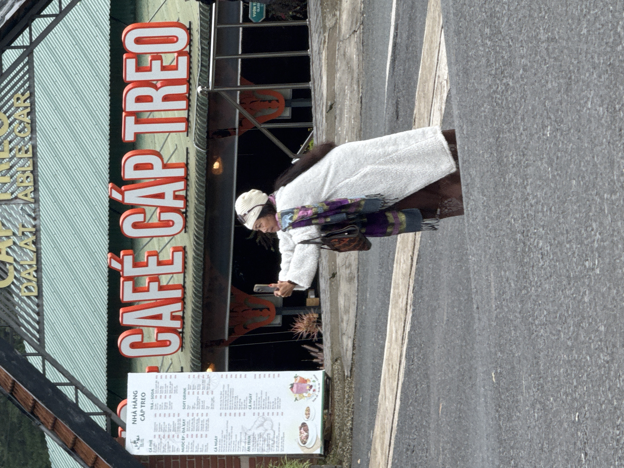 A woman is taking a selfie with her phone in front of a cafe called "CAFÉ CÁP TREO" in Da Lat, Vietnam. The cafe has large, red, illuminated lettering on its facade. A menu is visible on the left side of the image, listing various food and drink items. The woman is wearing a white fluffy coat, a headscarf, and a patterned scarf. She is holding a dark-colored bag over her shoulder. The cafe is located near a street, with a gray asphalt road in the foreground. The background shows greenery and a building with a green, corrugated roof. The scene appears to be outdoors, possibly during the day, given the lighting.