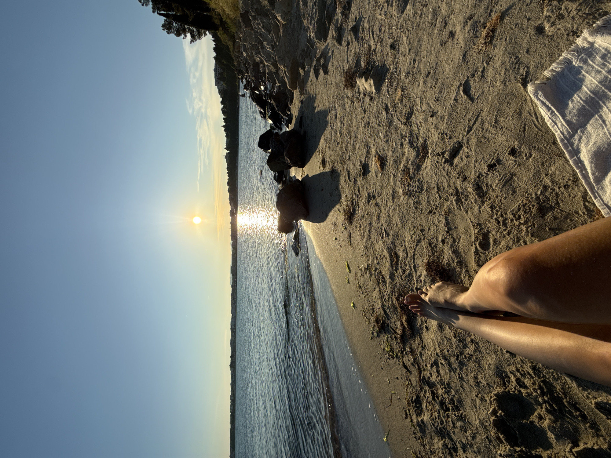 This media file captures a serene beach scene at sunset, with a person's legs visible in the foreground, resting on the sand. The individual is lying on a towel, which is white and has a fringed edge. Their legs are tanned and catch the warm, golden light of the setting sun, which is visible in the distance, low on the horizon, casting a shimmering reflection on the calm water.
The beach itself is sandy, with scattered rocks and some dark seaweed along the waterline. Beyond the water, a tree-lined shore is visible, with hints of buildings in the far distance, suggesting a coastal town or resort. The sky is a clear, pale blue, transitioning to a warmer, yellowish hue near the horizon where the sun is setting. The overall atmosphere is one of tranquility and relaxation, a perfect moment to enjoy the end of the day by the sea. The time of day is clearly sunset, indicated by the low angle of the sun and its warm light. The weather appears to be clear and pleasant. No visible text is present in the image.