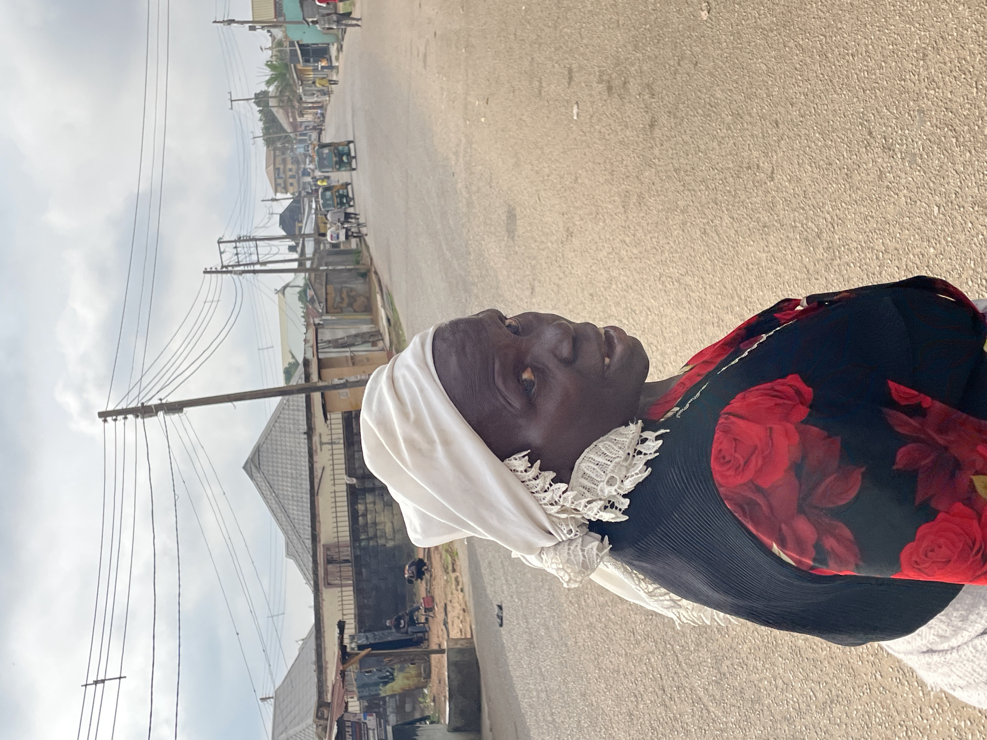 An elderly woman with dark skin and a white headscarf stands on a paved road in Jos, Nigeria. She wears a black and red floral-patterned top. The background includes buildings, power lines, and other individuals on the street, suggesting a populated urban environment. The light and shadows indicate it is daytime.