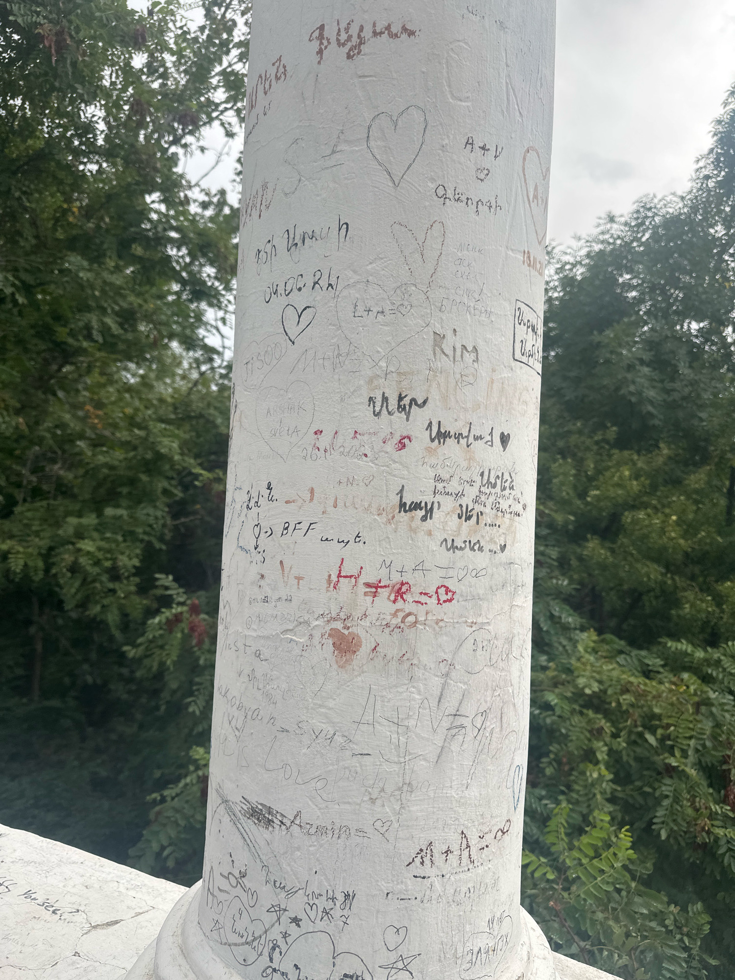 The image depicts a white stone pillar covered in numerous inscriptions, names, dates, and drawings, predominantly hearts. The pillar is located outdoors, with lush green trees and foliage visible in the background, suggesting a park or natural setting. The inscriptions appear to be in various languages, including Armenian and English. Some visible text includes "04.06.24", "BFF", "Love", "Armen", and what appears to be Armenian script. The pillar is part of a larger structure, with a white stone railing visible at the bottom left of the frame. This pillar is located in the vicinity of the "Mher Mkrtchyan Museum Theater" in Gyumri, Armenia, a known landmark where visitors often leave inscriptions. The overcast sky suggests a temperate climate.