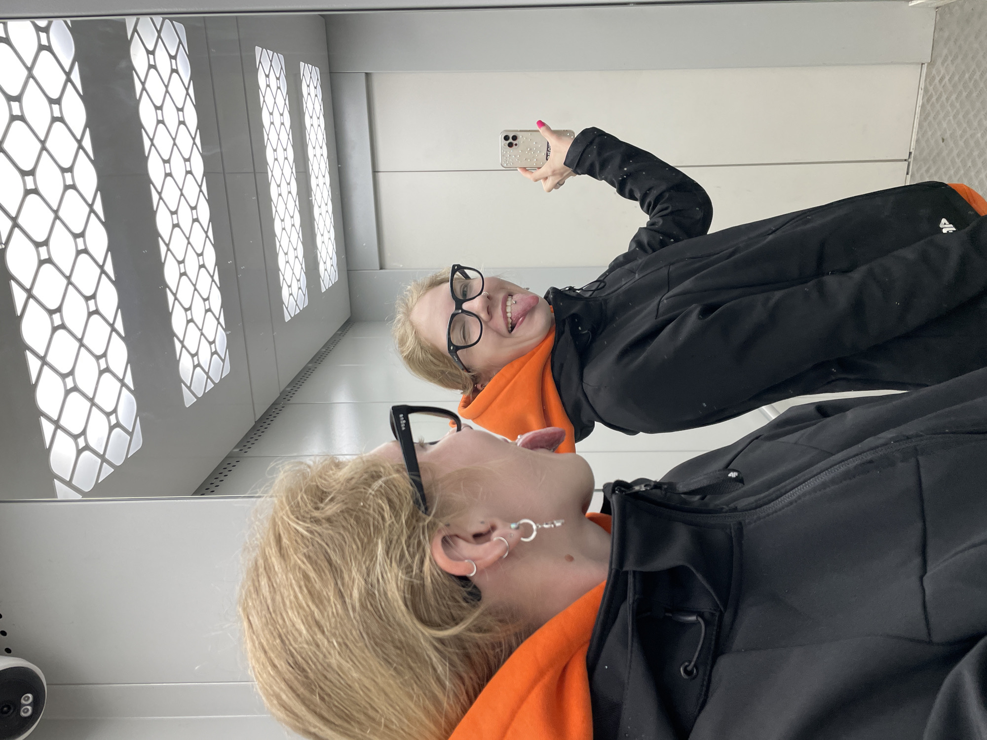 A woman is taking a selfie in a mirror inside a room resembling an elevator. The woman has blonde hair, glasses, and an orange top under a black jacket. She is sticking her tongue out at the camera. Her reflection is visible in the mirror. The interior is a white or light-colored room with patterned lighting fixtures. The location is presumed to be Izhevsk, Russia.