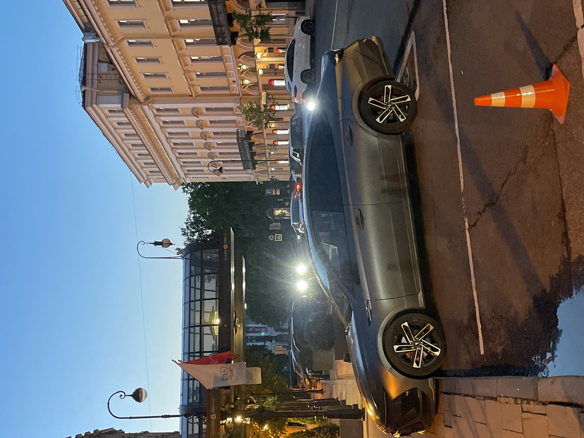The image depicts a city street scene during twilight or early evening in Saint Petersburg, Russia. The sky is a clear, deep blue, indicating the transition from dusk to night.
In the foreground, a dark grey luxury sport sedan, possibly a Porsche, is prominently parked on the right side of the asphalt road. Its headlights are on, creating bright lens flares. The car features distinctive multi-spoke wheels with a black and polished silver design. An orange traffic cone with white reflective stripes stands on the asphalt next to the car. A large, reflective puddle on the road surface mirrors the car and ambient lights. The road has white markings, and it transitions to a stone-paved sidewalk.
To the left, a multi-story classical-style building with a light-colored facade and numerous illuminated windows lines the street, showcasing ornate architectural details. Further down the street, other vehicles, including a white car, are parked.
On the right, a modern building with a glass facade is visible, behind which trees are silhouetted. Several streetlights with decorative, curved arms illuminate the area. A white banner with the number "150" and a logo can be seen. A "P" parking sign is also visible in the distance. The scene is characterized by the blend of natural low light and strong artificial illumination from car headlights, streetlights, and building interiors. The overall impression is an active urban environment at night.
