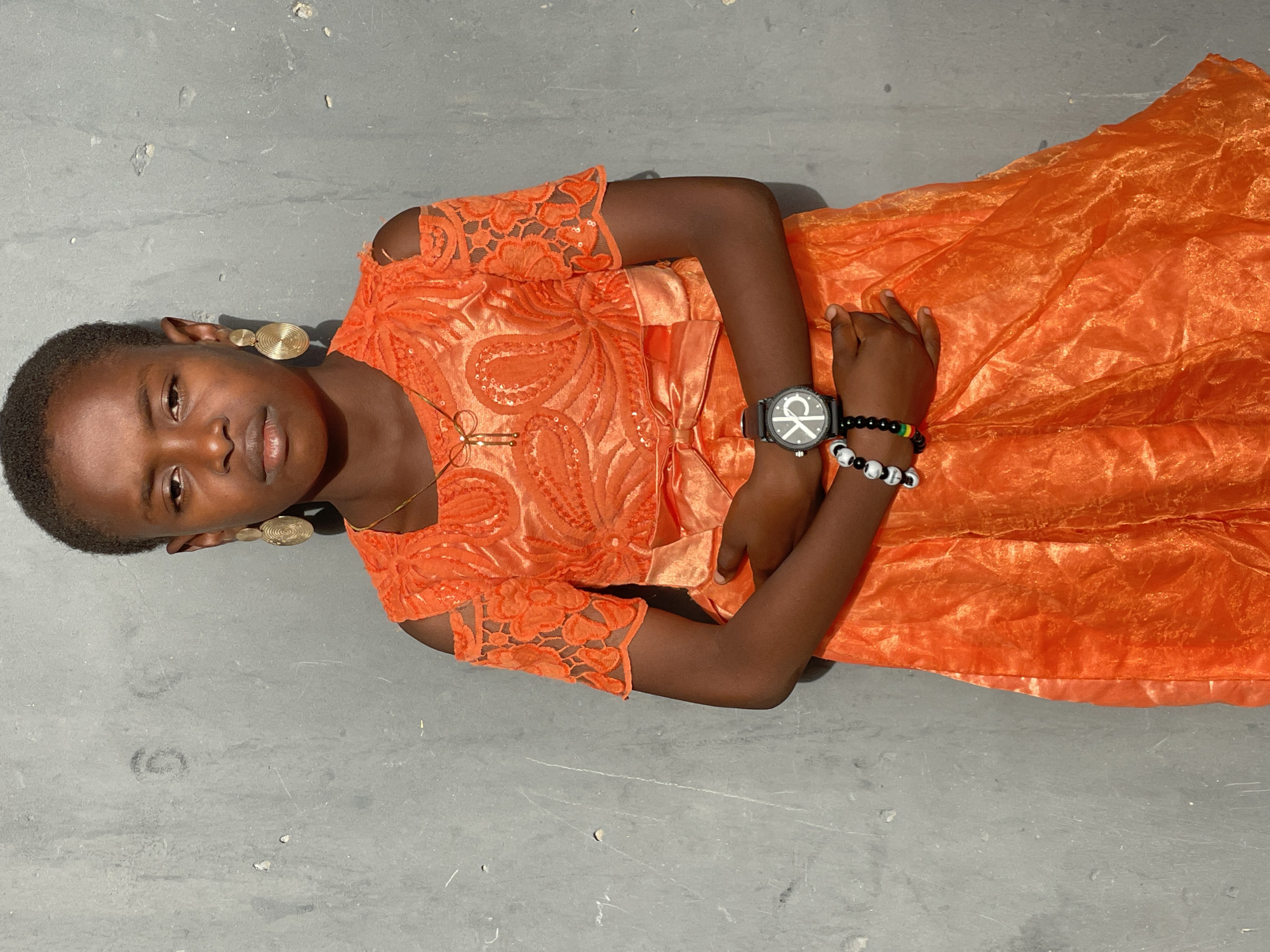 A young person, likely female, is depicted in a portrait-style photograph, lying on a light grey, somewhat textured concrete surface. They have dark skin and short, dark hair, and are looking directly at the camera with a calm, neutral expression.
The person is wearing a vibrant orange dress. The bodice and short, lace sleeves feature intricate, embroidered patterns, while the skirt is made of a flowing, sheer, and crinkled orange fabric. Notable accessories include large, golden, circular drop earrings with a spiral design, and a delicate gold chain around their neck with a small cross pendant. On their left wrist, they wear a black watch with a prominent white "CK" logo on its face. Adjacent to the watch are two beaded bracelets: one with black beads interspersed with small red, yellow, and green beads, and another featuring black and white patterned beads. Their arms are folded across their chest.
The background is entirely the grey ground, showing some minor scuffs and debris. The lighting appears bright and even, typical of daylight conditions. This scene is set in Badagry, Nigeria.