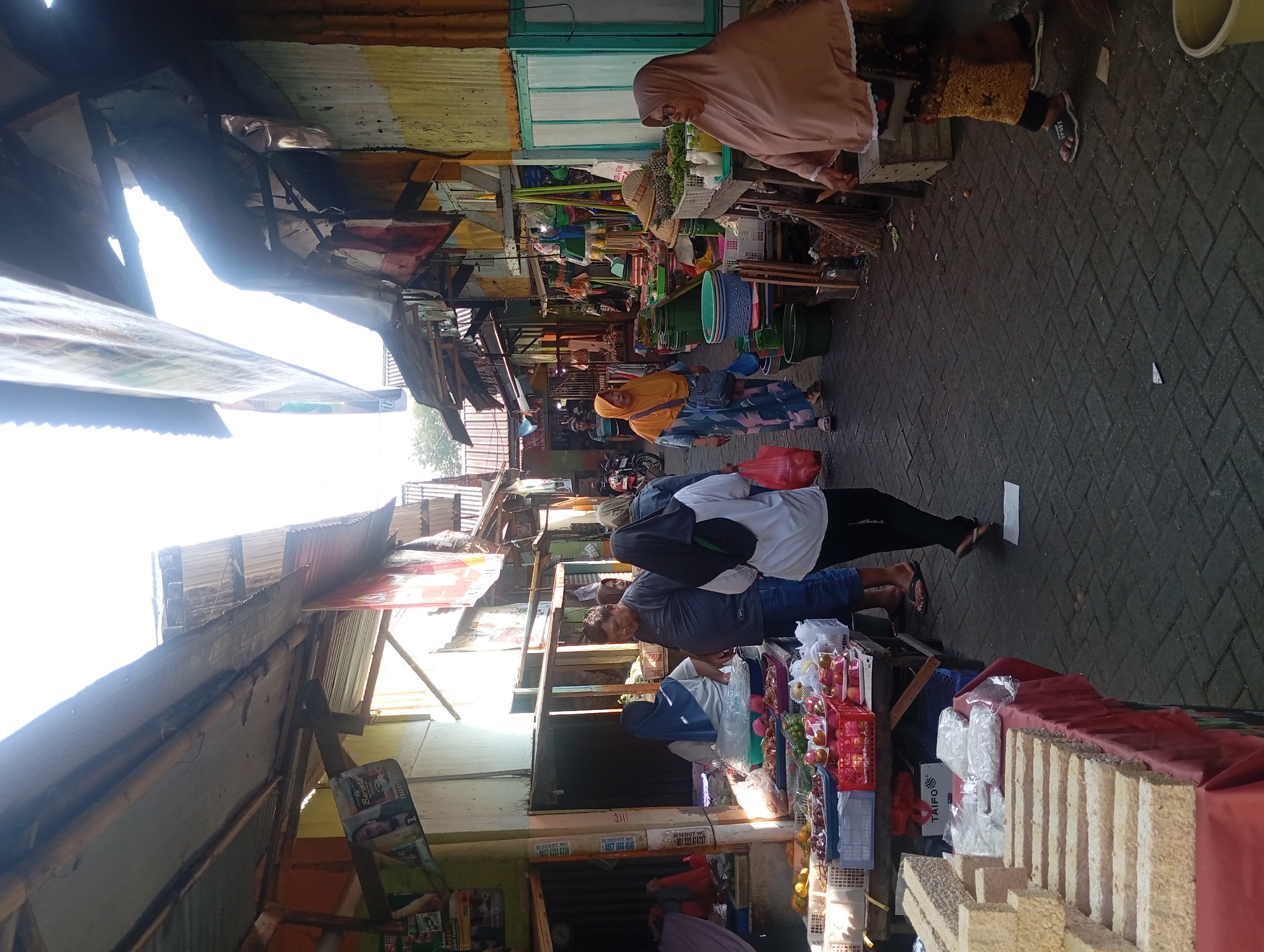 The image captures a bustling outdoor market scene in Kepulungan, Indonesia, bathed in bright sunlight that creates strong contrasts and shadows. The market appears to be situated along a paved pathway lined with stalls constructed from various materials, including corrugated metal, wood, and possibly tarps.
On the left side of the path, a stall displays an array of fresh produce, including what appears to be red apples, oranges, and possibly grapes, arranged in white and red plastic crates. Next to the produce, stacked rectangular blocks of what could be tempeh, a fermented soybean product, are prominently displayed on a table covered with a red cloth. Further down this stall, items wrapped in plastic bags are visible.
The central area of the image shows several people walking through the market. A woman in a blue hijab is behind a stall on the left, and a man in a gray t-shirt and blue jeans is interacting with someone near the produce. To the right of the man, a woman in a white shirt and black pants is walking away from the viewer, carrying a red plastic bag.
On the right side of the path, more stalls are set up, displaying a variety of goods. A woman wearing a peach-colored hijab and a patterned skirt stands near a stall laden with vegetables, possibly lettuce. Other stalls show plastic containers of various sizes, baskets, and what might be cleaning supplies. A straw hat is visible on one of the tables.
The architecture of the market is characterized by makeshift roofing structures, many of which appear to be made of corrugated metal, supported by wooden beams. These roofs create a covered walkway, but sunlight streams through gaps, illuminating the scene. The overall impression is one of a lively, everyday marketplace where commerce and daily life intersect.