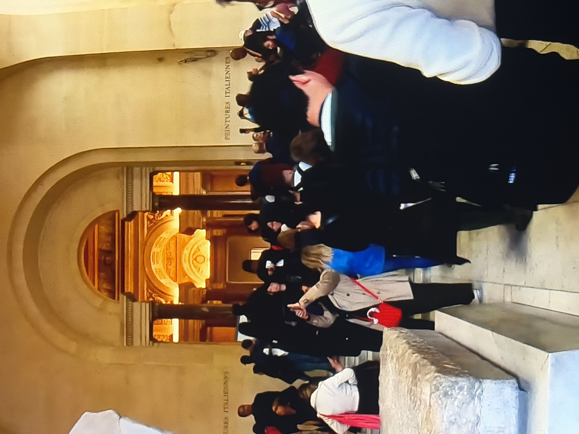 The image captures a group of people in what appears to be a museum or gallery. The architecture features grand arches and ornate detailing, suggesting a historical or culturally significant location. Two signs on the wall read "PEINTURES ITALIENNES" (Italian Paintings) and "TURES ITALIENNES" (likely "Peintures Italiennes" as well, with the first part cut off).
In the foreground, a large stone sculpture or display is visible on the left. Several individuals are scattered throughout the scene. On the left, a person in a white jacket with a pink scarf is standing, facing away from the camera. To their right, a person in a beige trench coat is holding a red bag and has their hand up, seemingly taking a photo or video. A person in a bright blue top is standing next to them.
The background shows a crowd of people, some silhouetted against the illuminated opening of a hallway or another section of the museum. The lighting is warm and inviting, emanating from the architectural elements and some unseen light sources. The overall atmosphere is one of a busy public space, with people engaging with the exhibits and the environment.