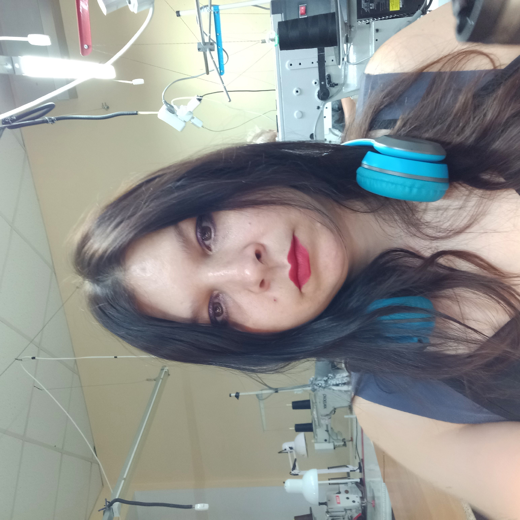 The image shows a close-up, frontal selfie of a woman with long, dark hair and blue headphones around her neck. She is wearing dark clothing and has red lipstick and purple eyeshadow. The background appears to be a workshop or factory setting, with sewing machines and overhead lighting visible. The room has light-colored walls and a suspended ceiling with fluorescent lights. There is no specific indication of the location within Kropyvnytskyi, Ukraine.