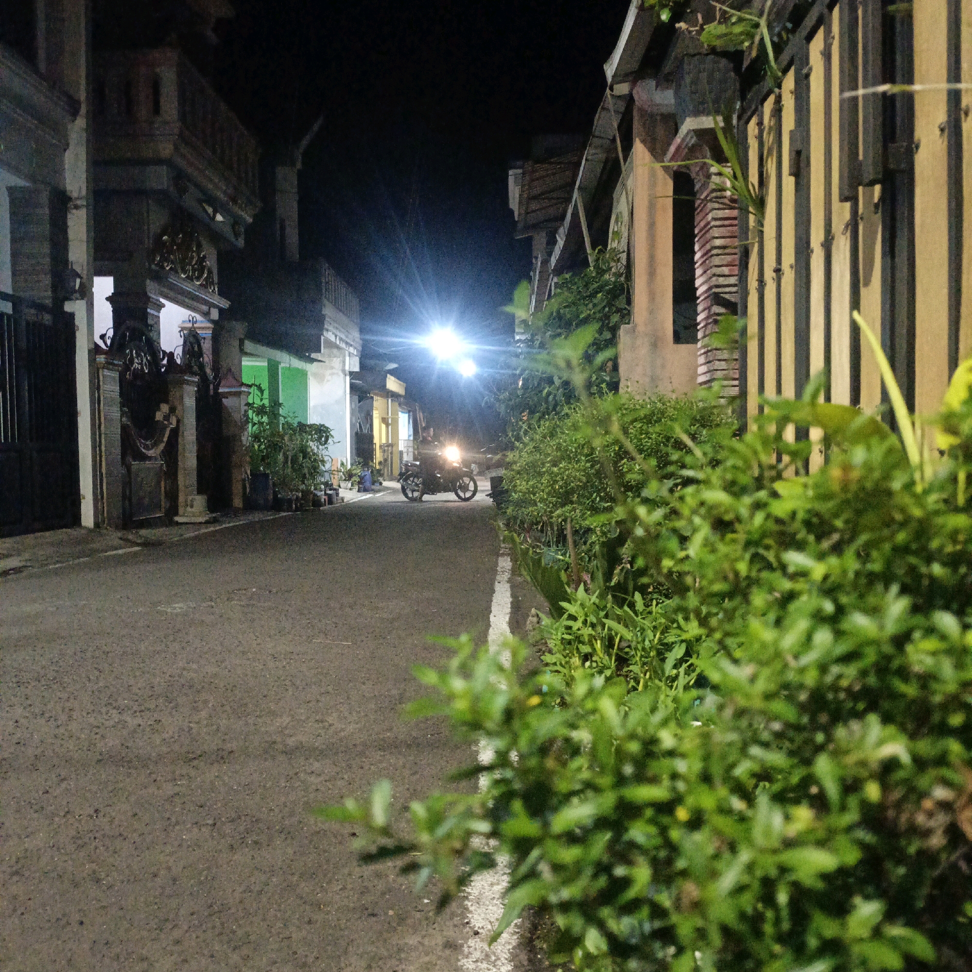 A motorcycle with its headlight on is parked on the right side of a narrow, paved street at night in Salatiga, Indonesia. The street is flanked by buildings on both sides. On the left, there are buildings with ornate architectural details, possibly houses or shops, with some gates and fences visible. On the right, a building with a yellowish-brown facade and vertical dark wooden planks is prominent, with lush green foliage in the foreground obscuring some of the building. The street has a white line painted on the right side. In the background, a bright light source, likely a streetlamp, illuminates the scene, creating a lens flare effect. The overall atmosphere is one of a quiet residential or commercial street at night.