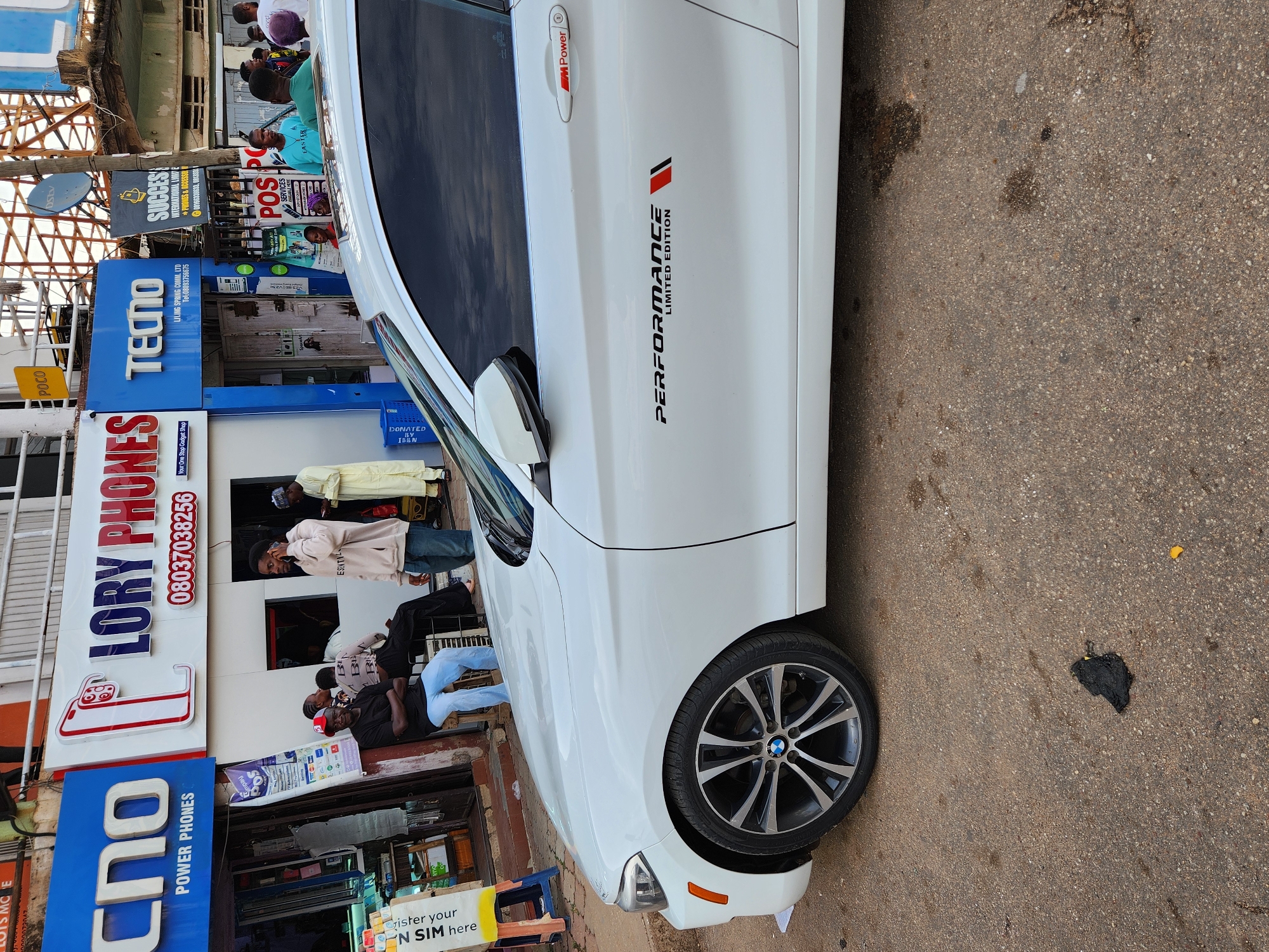 Here is a detailed description of the image:
**Scene Description:**
The image captures a street scene in an urban environment, likely in an African city. A white BMW car, identified by the "PERFORMANCE LIMITED EDITION" and "M Power" markings, is parked on the side of a paved road. There's a dark, tar-like stain on the pavement near the car. The background consists of several storefronts.
**Objects:**
* **Vehicles:** White BMW car.
* **Signage:** Signs for "LORY PHONES", "TECTO", "SUCCESS INTERNATIONAL LIMITED", "Cho" and a sign referencing N SIM registration.
* **Infrastructure:** Overhead electrical wires or cables, building structures, and a satellite dish are visible.
* **Miscellaneous:** Displayed phones, boxes, a donation crate labeled "DONATED BY IBBN"
**People:**
Several people are present, some standing outside the storefronts. Details like gender, clothing are visible.
**Location:**
The location is identified by the signage of local businesses "LORY PHONES", "TECTO", and "SUCCESS INTERNATIONAL LIMITED". It is likely in an African city, though the exact city remains unknown.