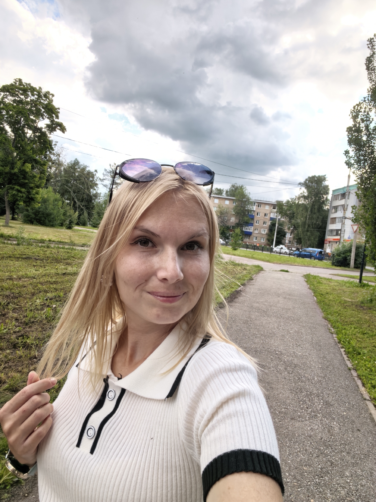 A blonde woman is taking a selfie outdoors on a cloudy day, standing on a paved walkway in a city. She is wearing a white polo shirt with black trim and buttons, and her sunglasses are perched on top of her head. She is holding a strand of her hair with her left hand. Behind her, to the left, is a grassy area with trees, and to the right, there is a road with cars and apartment buildings. The scene appears to be in an urban residential area, possibly in Sterlitamak, Russia, given the architectural style of the buildings and the presence of a road and walkway typical of Russian cities.