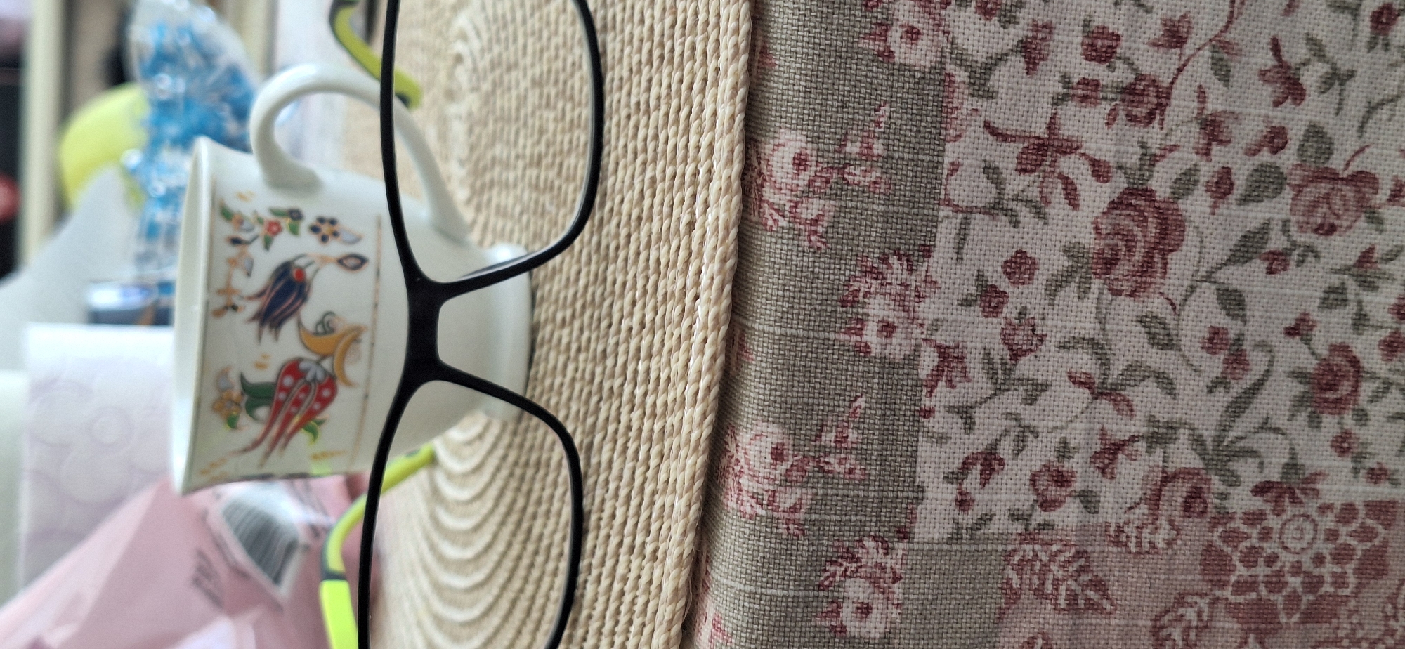 The image shows a close-up of several items placed against a backdrop with a floral pattern. In the foreground, a pair of black-framed glasses is positioned to the left. Behind the glasses, there's a white teacup with colorful decorative motifs, including birds and floral elements. The teacup appears to be hanging, possibly from the glasses or another unseen object.
To the right of the glasses and teacup, a woven beige mat or surface adds texture to the composition. The backdrop consists of a patterned fabric or wallpaper with a floral design featuring roses in various shades of red and green.
Based on the provided information, it is impossible to determine the specific location of the scene in the city of Blagoevgrad, Bulgaria, as the image lacks contextual clues regarding the setting.