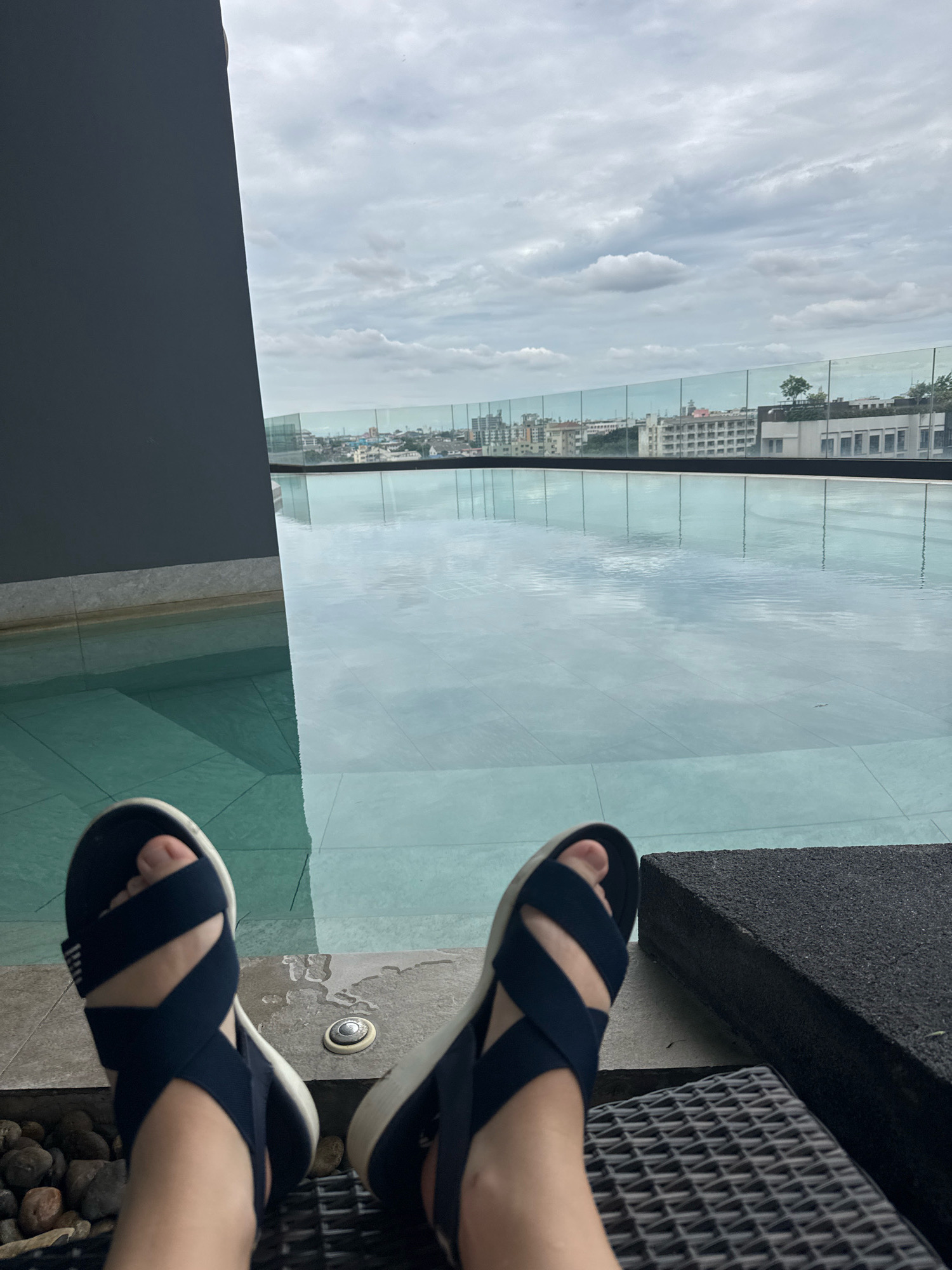 The image shows a person's feet wearing navy blue sandals with white soles, resting on a dark woven chair. The feet are positioned in the foreground, with a shallow swimming pool in the middle ground. The pool's surface reflects the cloudy sky and buildings in the background. Beyond the pool, a glass railing separates it from a cityscape. The buildings appear to be a mix of residential and commercial structures. The sky is overcast with scattered white clouds. A dark gray wall is visible on the left side of the image, suggesting an architectural element of the location. The overall scene appears to be a rooftop or elevated area with a pool overlooking the city. The location is Samrong Nuea Subdistrict Municipality, Thailand.