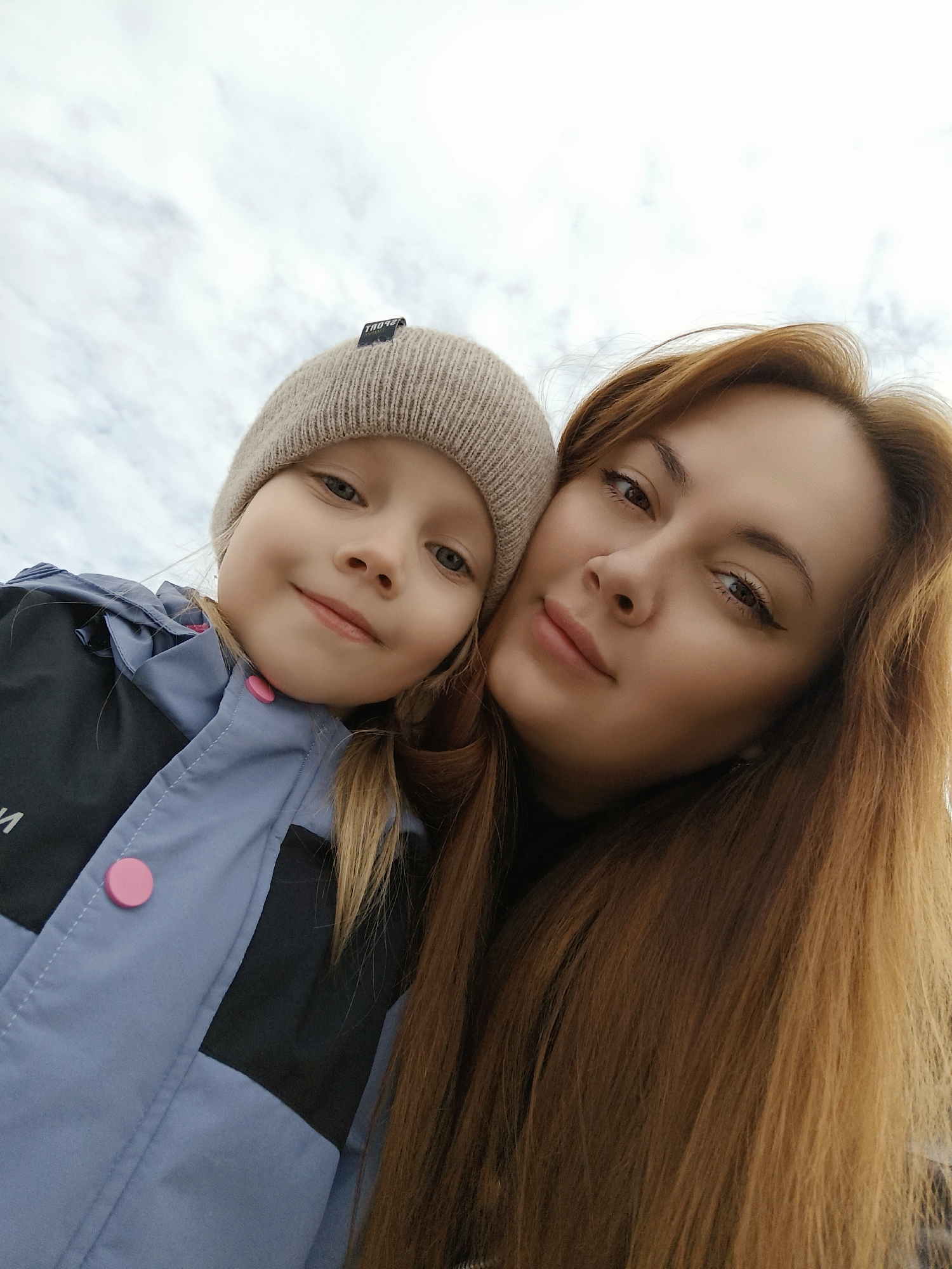 The image features two individuals, an adult female and a young girl, in a close-up, low-angle shot against an overcast sky. The young girl, estimated to be between 5 and 8 years old, is positioned on the left. She wears a light beige knit beanie with a small dark label on its front cuff and a light blue-grey and black panelled jacket with prominent round pink buttons on the front closure. Her blonde hair is partially visible, and she exhibits a smiling expression, looking directly at the camera. The adult female, positioned on the right, has long light brown hair, visible eye makeup, and a neutral to slightly smiling expression, also looking directly at the camera. Her head is close to the girl's, indicating a close relationship. The background is uniformly composed of diffuse, cloudy sky, offering no discernible landmarks or contextual information regarding a specific location within the city of Chaikovsky, Russia.