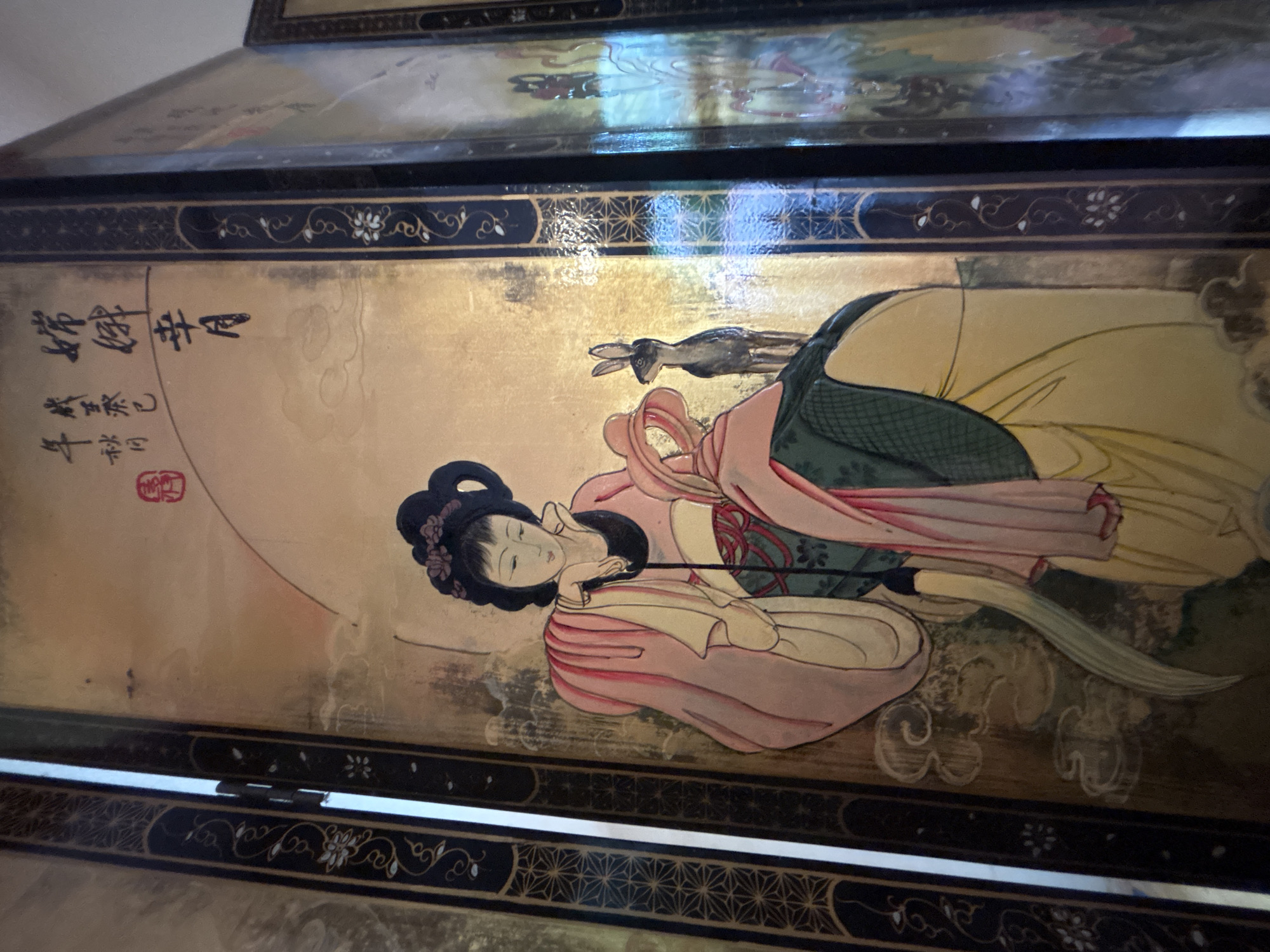 This is a close-up, eye-level, indoor shot of a segment of an antique, gold-leaf decorated folding screen with a painting of two figures and a rabbit. The screen is dark brown with ornate gold and black borders featuring floral and geometric patterns. The painting depicts two figures in traditional East Asian attire. The figure on the left is a woman with her hair styled in an updo, wearing a pink robe over a green garment. She is leaning on her hand, looking downwards. The figure on the right is partially visible, wearing a pink head covering and a green robe over a yellow garment, with their back to the viewer. A small, dark grey rabbit stands between the two figures, looking towards the left. Chinese calligraphy and a red seal are visible in the upper right quadrant of the screen. The background of the painting is gold with faint cloud-like motifs. The lighting suggests an indoor environment, possibly a home or gallery, with reflections on the screen's surface. The location of this screen in Chalong, Thailand is not discernible from the image itself.