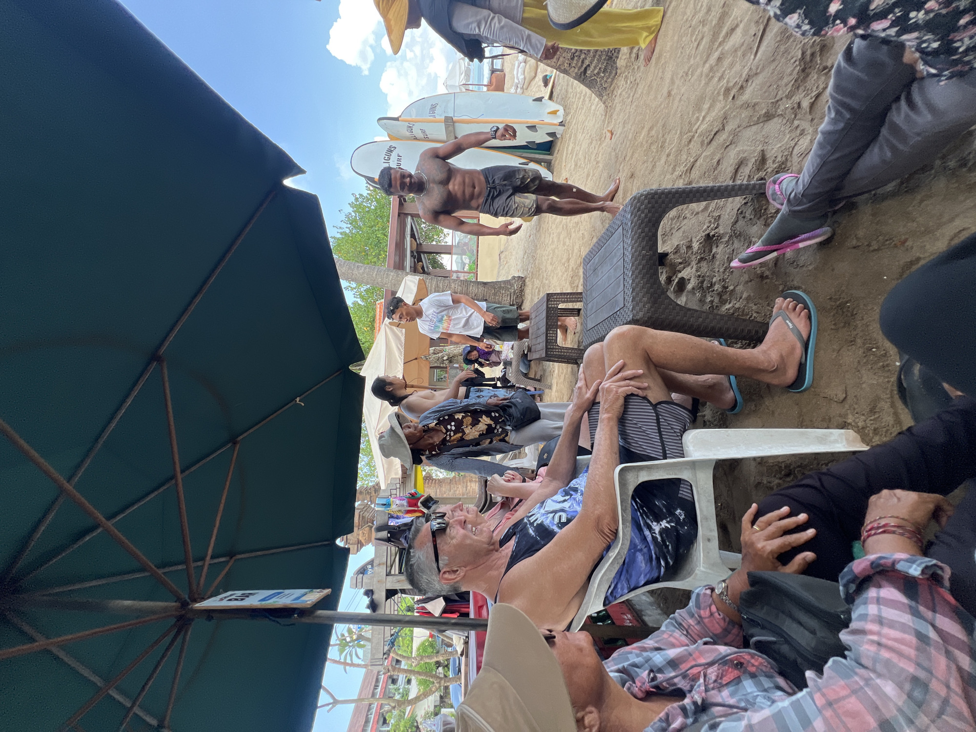 In Kuta, Indonesia, a group of people are gathered on a sandy beach beneath a large green umbrella. The scene appears to be a beachside cafe or resting area. In the foreground, a man and a woman wearing hats are seated, facing left. The man, with sunglasses and a black and blue tank top, sits in a white plastic chair. Next to him, a woman in a pink plaid shirt is also seated. Further back, another man with sunglasses and a black tank top is visible, and behind him, a woman in a hat and patterned shirt is standing. A group of surfboards with "LIFIGUNS SURF" written on them are leaning against a palm tree on the right side of the frame. The ocean and sky are visible in the background, with a few buildings and trees also present. The overall atmosphere suggests a relaxed beach outing.