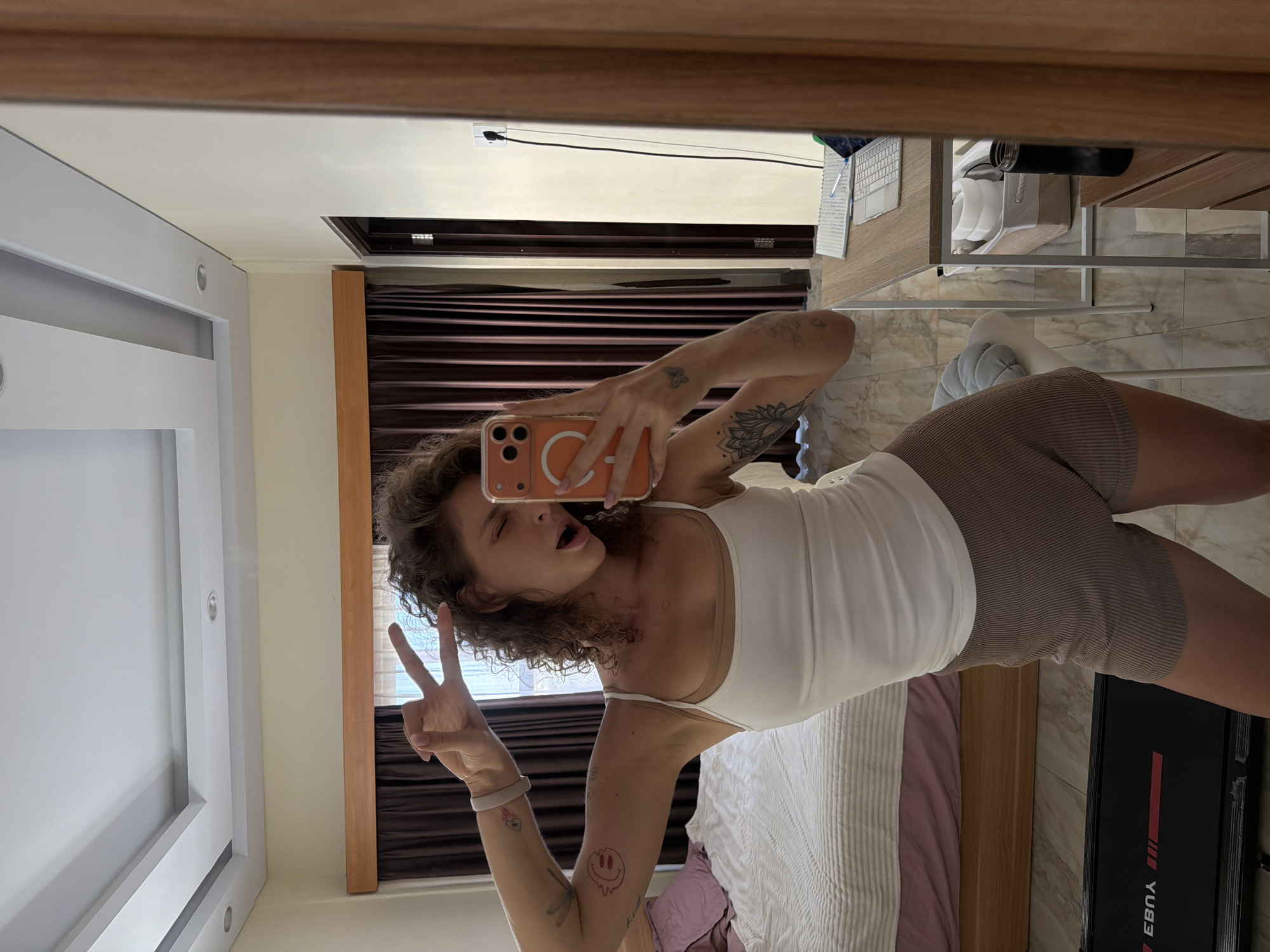 A woman with curly brown hair is taking a mirror selfie in a room. She is wearing a white tank top and brown shorts. Her left arm is raised, and she is making a peace sign with her fingers. She has several tattoos on her arms, including a lotus flower, a butterfly, and a smiley face. In the background, there is a bed with pink and white linens, dark brown curtains, and a desk with a laptop and notebook. A treadmill is visible in the foreground. The room appears to be in a hotel or rental property in North Kuta, Indonesia, indicated by the architecture and furnishings.