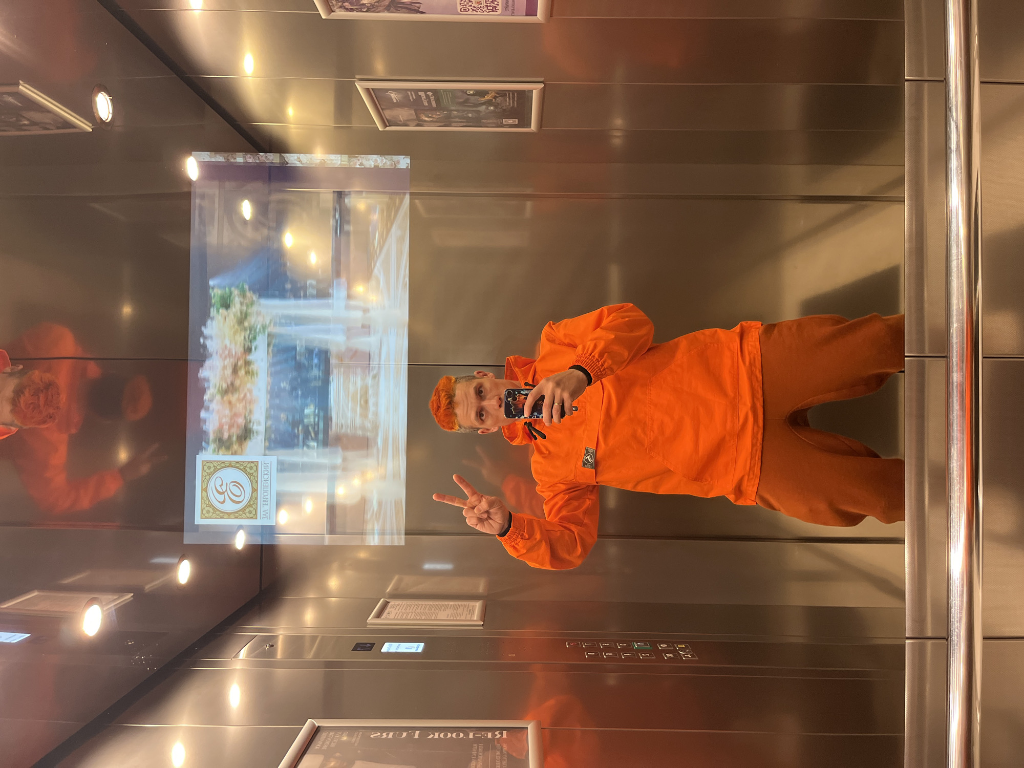 A man wearing an orange sweatshirt and pants stands inside an elevator, holding a phone and making a peace sign. The elevator has brushed metal walls, ceiling lights, a vertical control panel with buttons, and a screen showing a scene of a fountain and floral arrangement. The screen also has a logo with Cyrillic text that identifies the location as "ZAO Evropeiskiy" (ЗАО Европейский). Based on this information, the man is most likely in an elevator at the European Shopping Centre, also known as Evropeiskiy Mall, in Moscow, Russia. There are framed posters at the top and bottom of the elevator. The man is reflecting in the elevator's mirrored wall.
