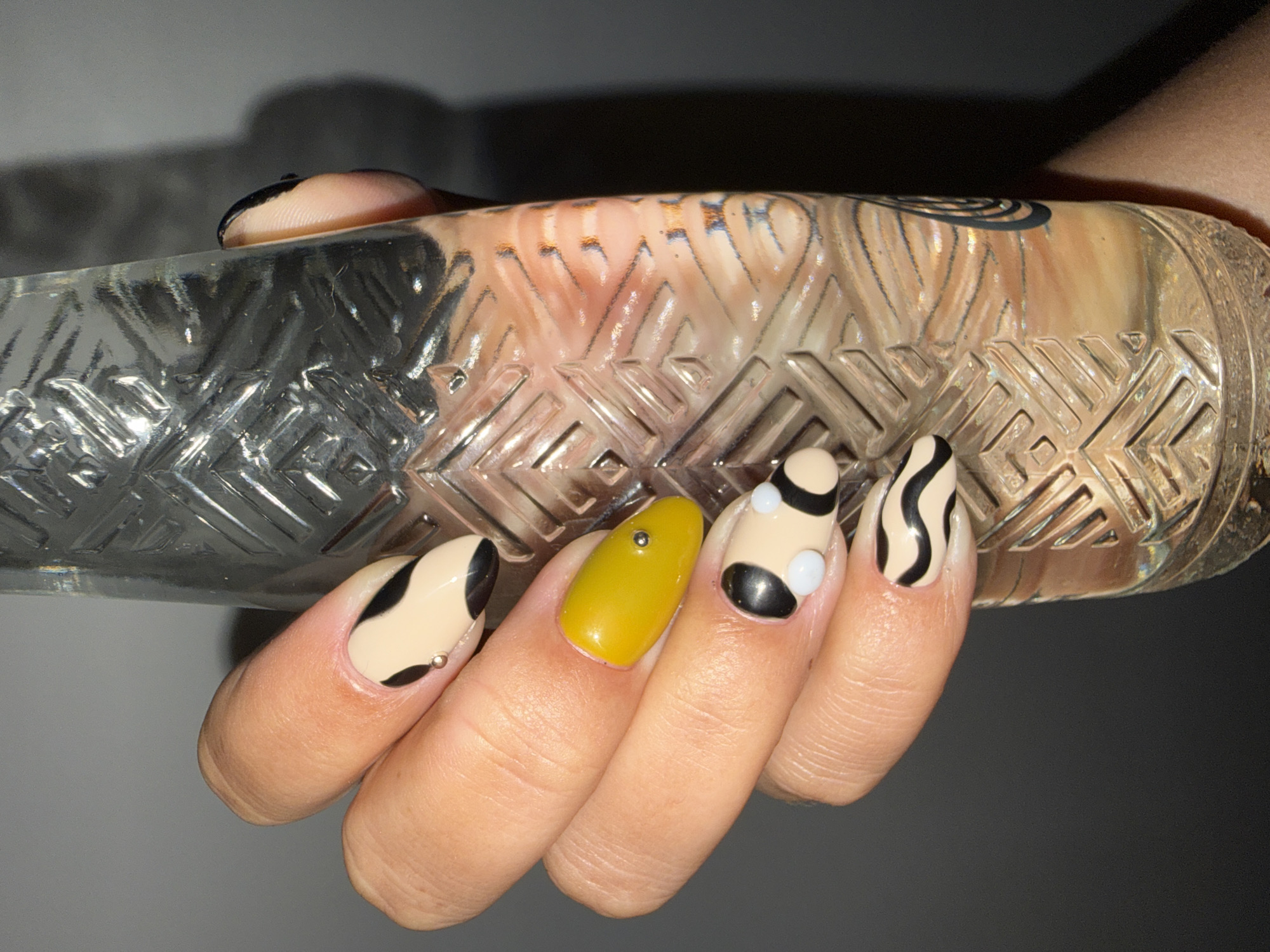 A hand is holding a clear glass bottle with a textured pattern. The hand has different nail art designs: one nail is yellow with a small silver bead, two nails have a tan base with black curved shapes, and one nail has black and tan zebra stripes. The background is a gradient from light gray to dark gray. North Kuta, Indonesia is not verifiable from the given information.