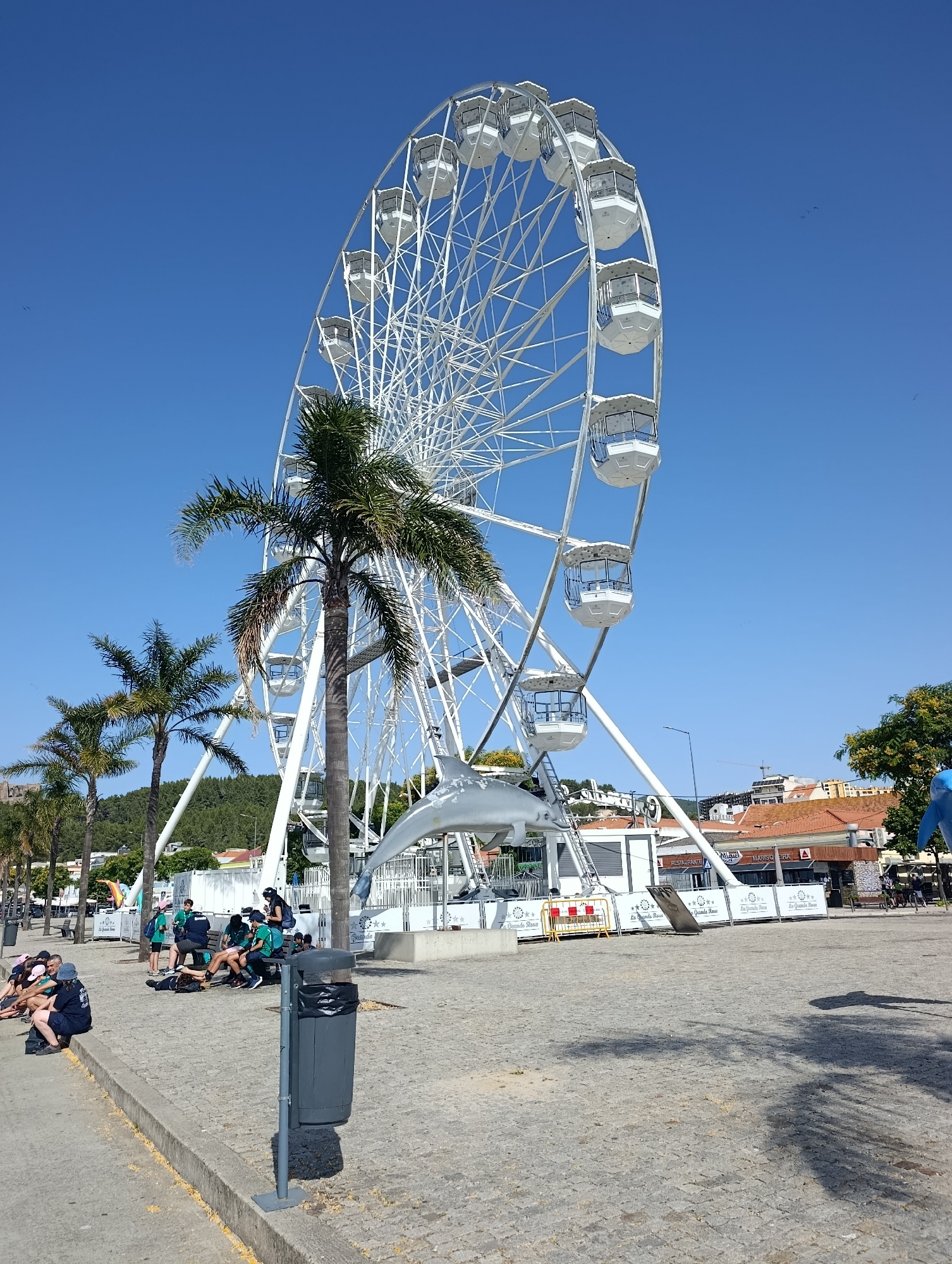 This media file captures a sunny day at a waterfront amusement area in Setúbal, Portugal. A prominent white Ferris wheel dominates the scene, with several of its enclosed cabins visible against a clear blue sky. In the foreground, several palm trees stand tall, casting shadows on the paved ground.
A group of people, appearing to be tourists or students, are relaxing or gathered near the base of the Ferris wheel. They are dressed casually, some in green t-shirts. Their postures suggest a relaxed atmosphere, with some sitting on the ground and others on benches. A dark gray trash can with a black bag stands near the group.
In the background, buildings with terracotta roofs are visible, suggesting a coastal town setting. The ground is paved with cobblestones. A metal sculpture resembling a dolphin is attached to the structure of the Ferris wheel. Fencing and signs are also visible around the attraction. The overall impression is one of a bright, pleasant day at a tourist destination.