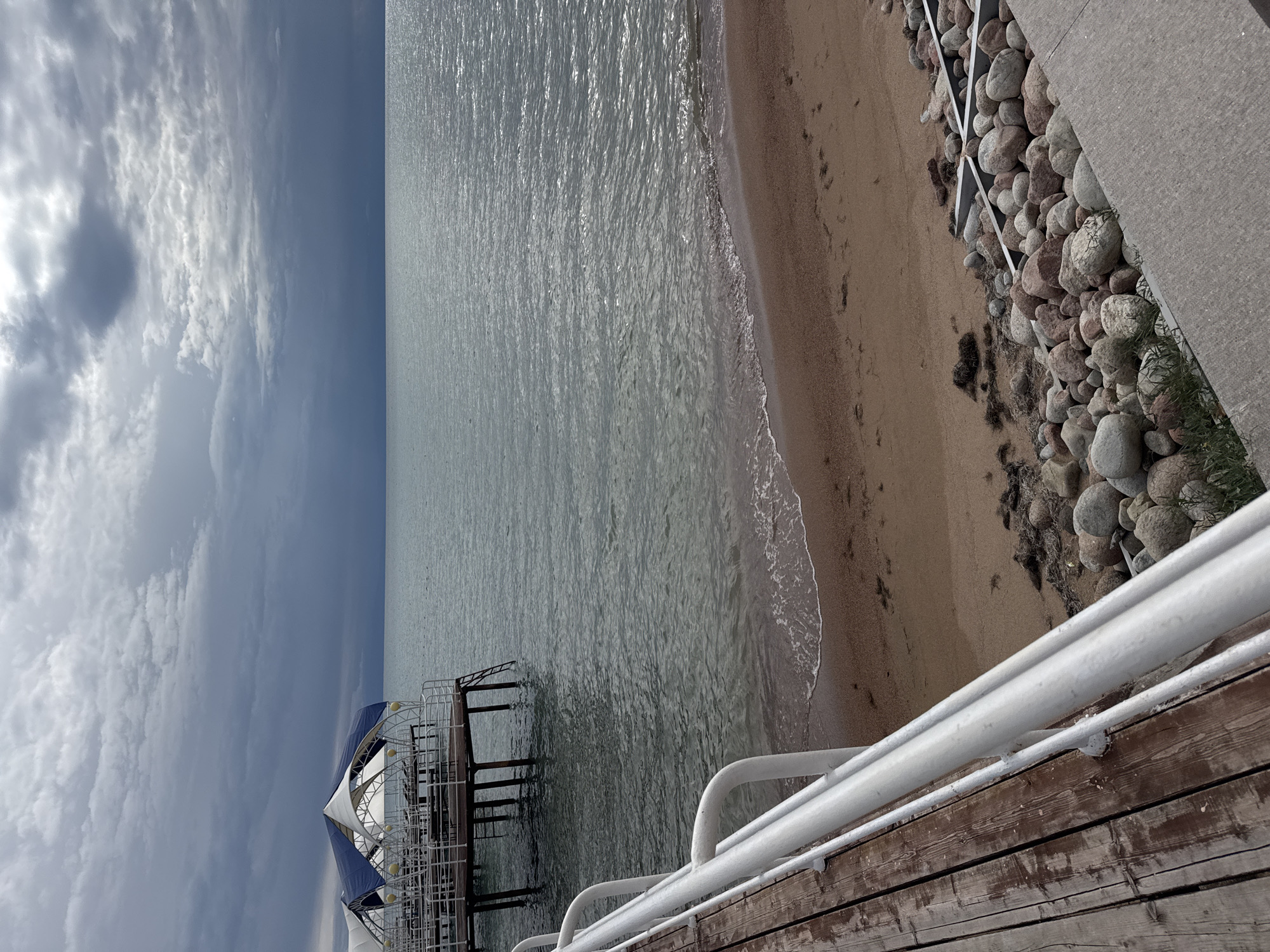 The image depicts a tranquil scene at the shore of Issyk-Kul Lake in Sary-Oy, Kyrgyzstan. A wooden pier with white railings extends into the calm, grey water. At the end of the pier, a structure with a blue and white canopy provides shade, suggesting a seating area or a small cafe. The beach is composed of fine sand, with a line of rounded stones bordering the water's edge. The sky is overcast with dramatic, dark clouds, hinting at impending rain. The overall atmosphere is serene and slightly somber due to the weather conditions.