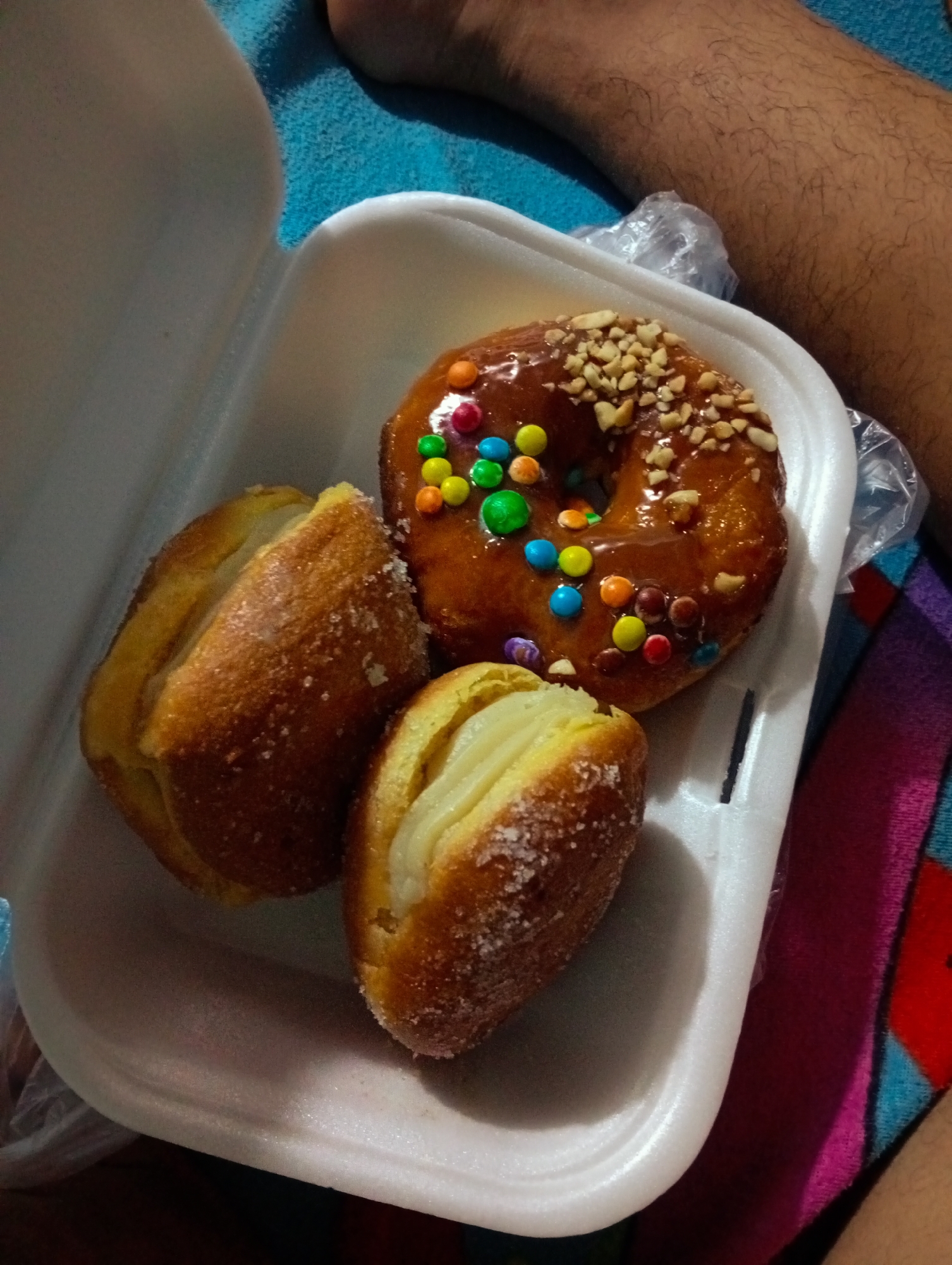 The image shows a white styrofoam container filled with three pastries. One is a doughnut, glazed with a brown coating and decorated with colorful candies and chopped nuts. The other two pastries are oblong, filled with cream, and coated in sugar. The container sits on a surface with a blue and magenta cloth. A person's leg is partially visible in the upper and right areas of the image. It is impossible to determine the location of the scene within Altagracia, Venezuela, based solely on this image.