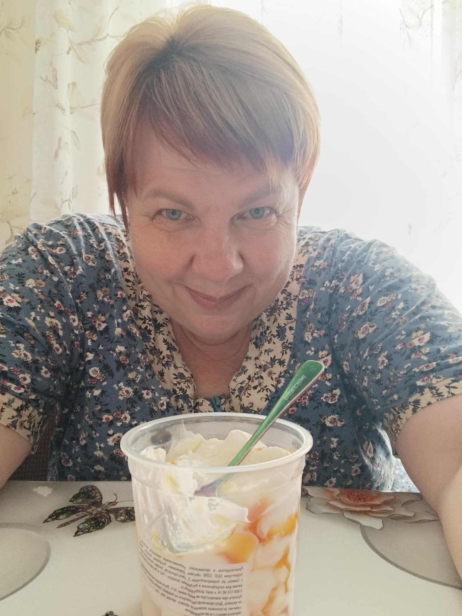 A woman with short blonde hair and blue eyes is looking at the camera and smiling. She is wearing a blue floral-print shirt. In front of her is a clear plastic cup filled with a creamy dessert, possibly ice cream or yogurt, with pieces of fruit. A green spoon is sticking out of the cup. The scene appears to be indoors, near a window with sheer curtains. The table the cup is on has a floral pattern. The overall lighting suggests it is daytime. Based on the provided context, the location is Svietlahorsk, Belarus.