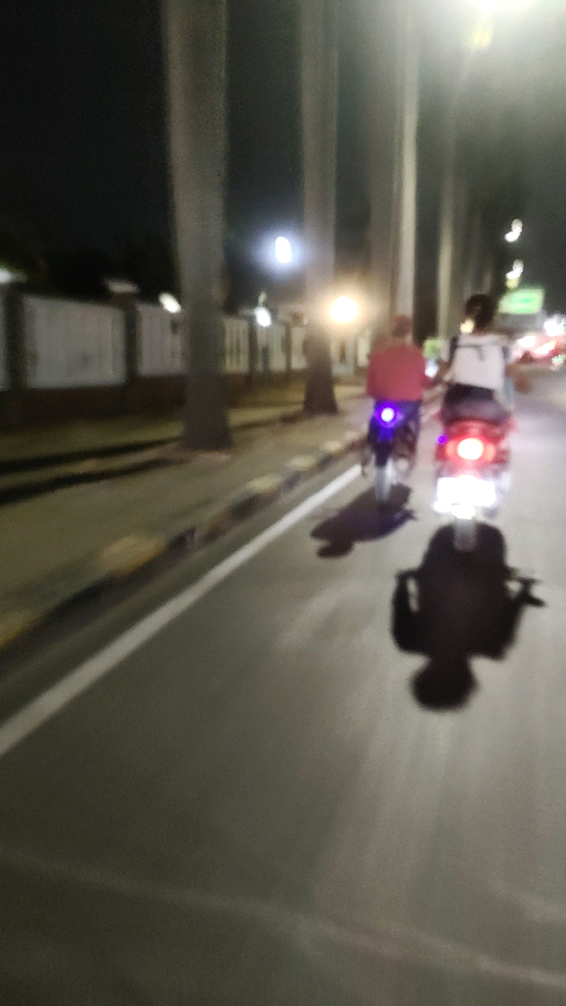 In Mekarmukti, Indonesia, at night, two individuals are riding motorcycles on a paved road. The motorcycle on the right has its brake lights illuminated. The rider on the right is wearing a white top. The rider on the left is wearing a red top. The scene is illuminated by streetlights and the headlights of the motorcycles. The road appears to be lined with trees and a fence. The image is blurry, suggesting motion blur.
