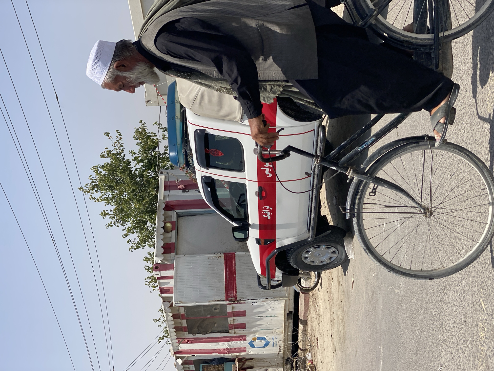 An elderly man wearing a white skullcap and a dark vest over a black shirt rides a bicycle. His grey beard is full and long. He is holding the handlebars of the bicycle, which is positioned next to a white vehicle with red stripes. The vehicle appears to be a service vehicle or ambulance, with some text in red on the side. The vehicle is parked on a paved surface, possibly a street. Behind the vehicle, there is a small, striped building with some trees and power lines visible against a clear, bright sky. The lighting suggests it is daytime. The overall scene is a snapshot of daily life in Kandahar, Afghanistan. The man's posture and the context suggest he is engaged in an everyday activity.