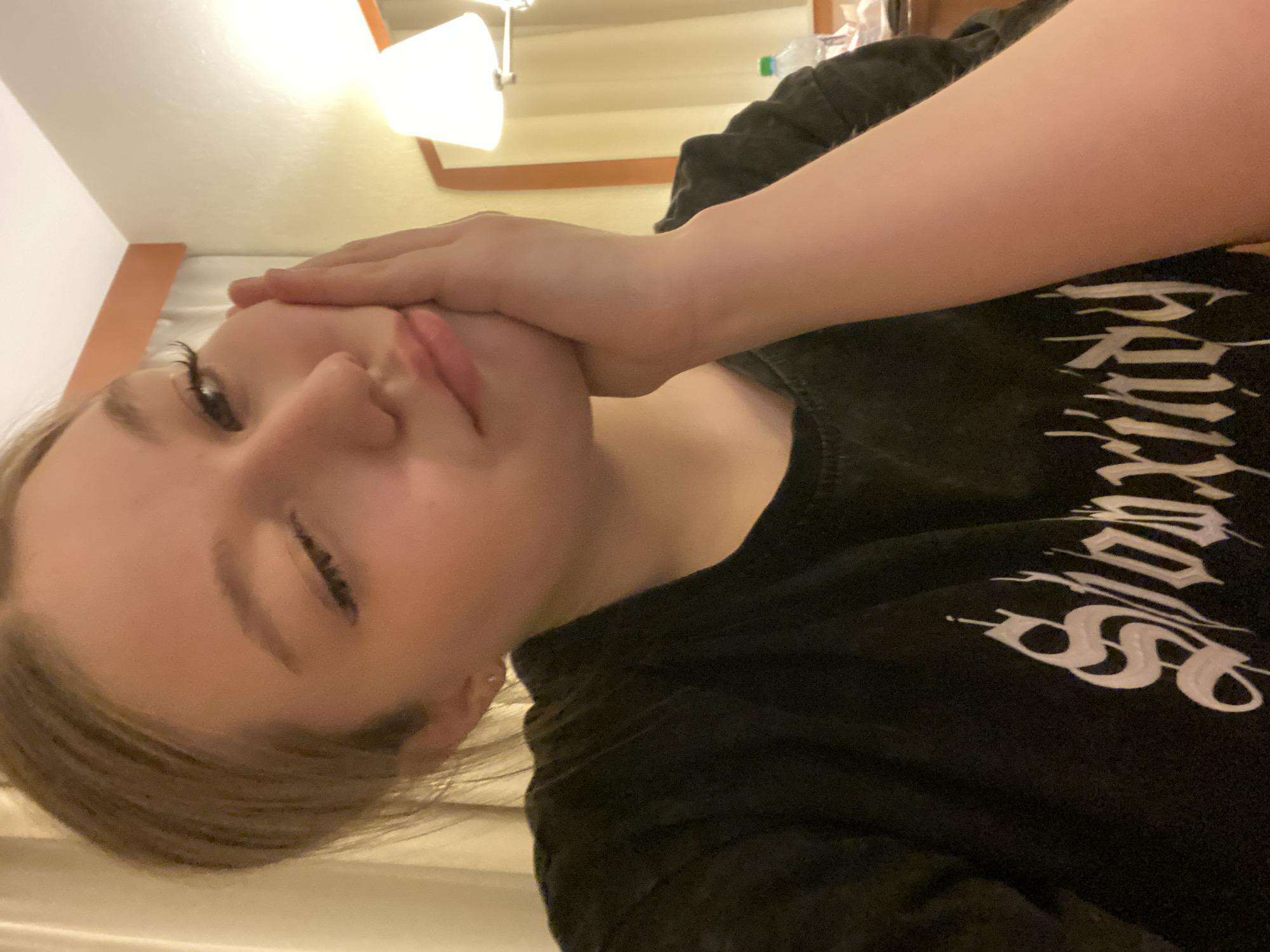 A person with light skin and light brown hair is pictured indoors. They are wearing a black T-shirt with white text. Their right hand is covering the bottom half of their face. There is a lamp visible in the upper left corner of the frame. The location is not identifiable as Saint Petersburg, Russia.