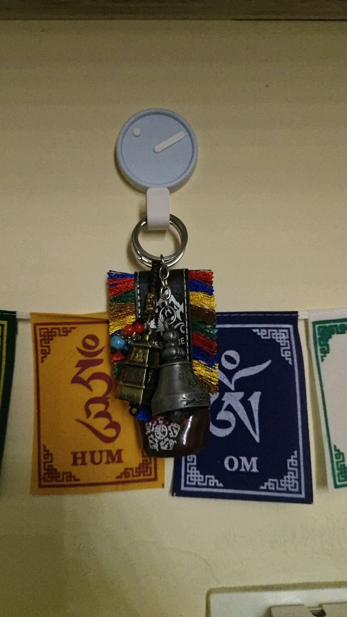 A pale yellow wall serves as the backdrop for a close-up, eye-level shot of a keychain hanging from a white, circular clock-like object. The keychain features a metal ring with a braided brown string attached, from which dangle several charms. These charms include a small, multi-tiered pagoda-like structure, a larger bell, and a dark, translucent object with what appears to be a white design etched onto it. Colorful beads in red and blue are also present on the string.
Below the keychain, a string of prayer flags stretches horizontally across the image. The prayer flags are made of fabric, with the closest ones being yellow and dark blue. The yellow flag has Tibetan script and the word "HUM" printed in dark red. The dark blue flag displays a white symbol and the letters "OM". The prayer flags are adorned with intricate white borders. The overall scene is well-lit, and the focus is sharp on the keychain and the prayer flags. No people are visible in the image. The image does not provide any specific information about the city of Chithera, India.