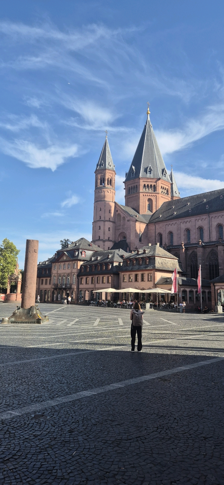 The image depicts a sunny day in Mainz, Germany, specifically in front of the Mainz Cathedral (Mainzer Dom). The cathedral's prominent reddish-brown stone structure with its tall spires and arched windows dominates the background. In the foreground, a person with a backpack stands in a large, open cobblestone square. The square features intricate geometric patterns formed by lighter colored stones on a darker cobblestone surface. To the left, a tall, weathered stone column stands next to a fountain-like structure. Behind the column and to the left of the cathedral, trees are visible. In the middle ground, a row of buildings with awnings and outdoor seating suggests a cafe or restaurant area. Two red flags with white lettering are prominently displayed on poles in front of one of the buildings. The sky is bright blue with wispy white clouds. The scene is captured from a slightly low angle, emphasizing the scale of the cathedral.