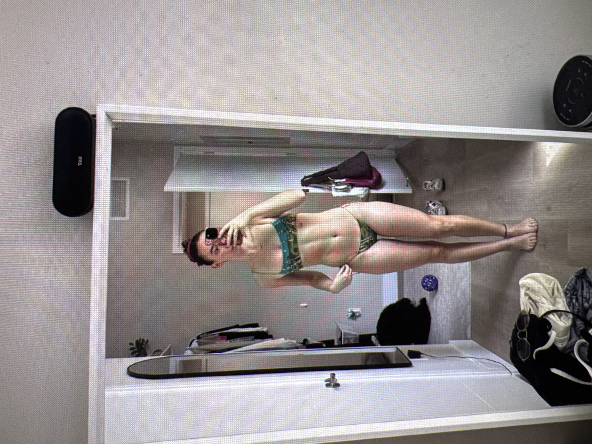 A young woman stands facing a mirror, taking a selfie. She is wearing a green and blue patterned bikini. Her hair is pulled up, and she is holding a cell phone in her right hand to capture the reflection. The background shows an indoor scene, possibly a bedroom or dressing area. There is a closet to her left with clothes hanging inside, a full-length mirror, and a speaker on top of the mirror frame. On the floor, there are shoes, a ball, and some clothing items. The overall lighting suggests it is daytime. The provided information about the city of Castelo Branco, Portugal, cannot be verified from the image alone.
