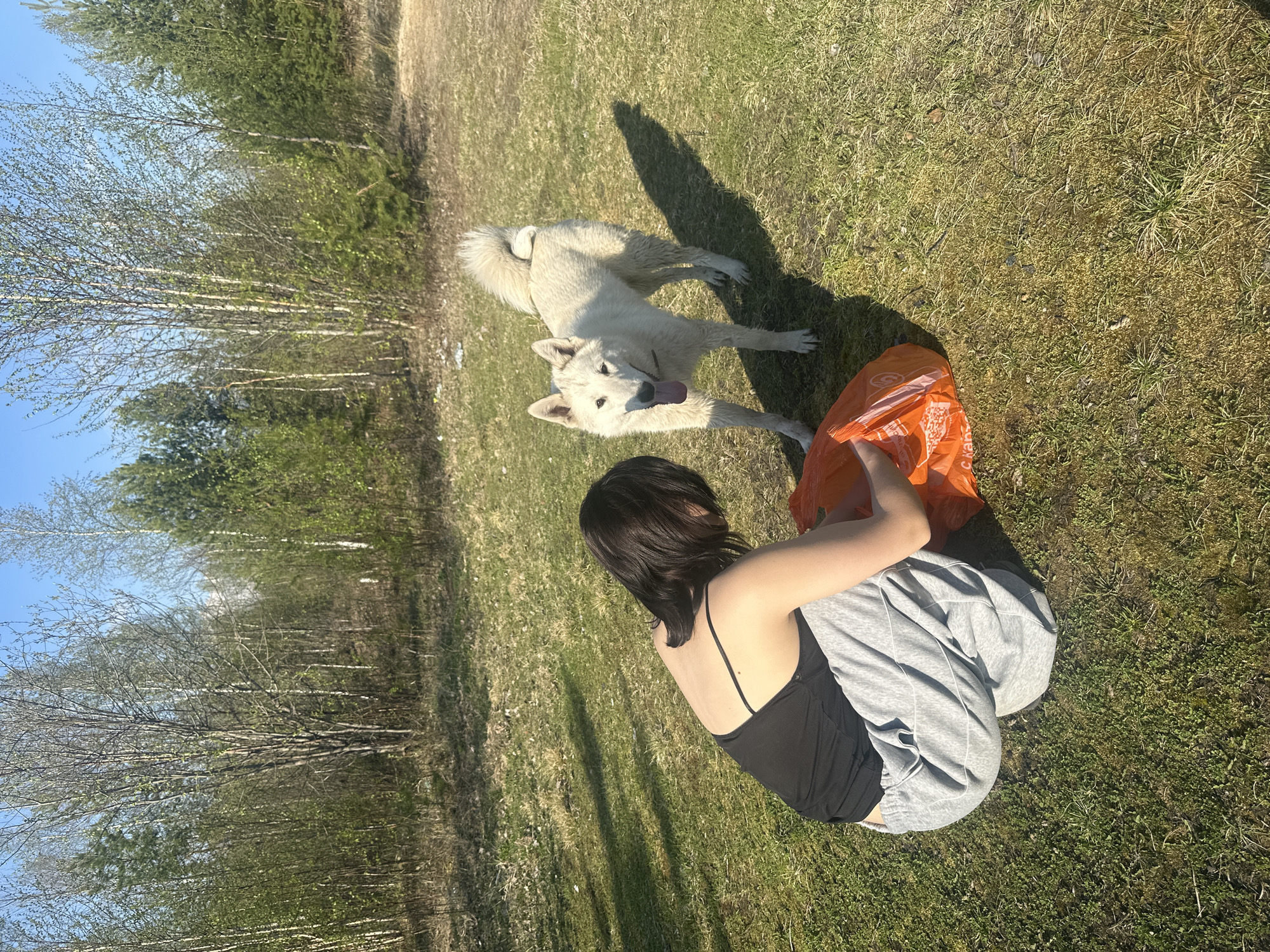 The image shows a woman sitting cross-legged on a grassy field. She is wearing a black tank top and gray sweatpants, and her back is facing the camera. An orange plastic bag is near her. A white dog is standing near her, with its tongue out. The field is partially shaded and there are trees in the background. The location is in the city of Vysokii, Russia.