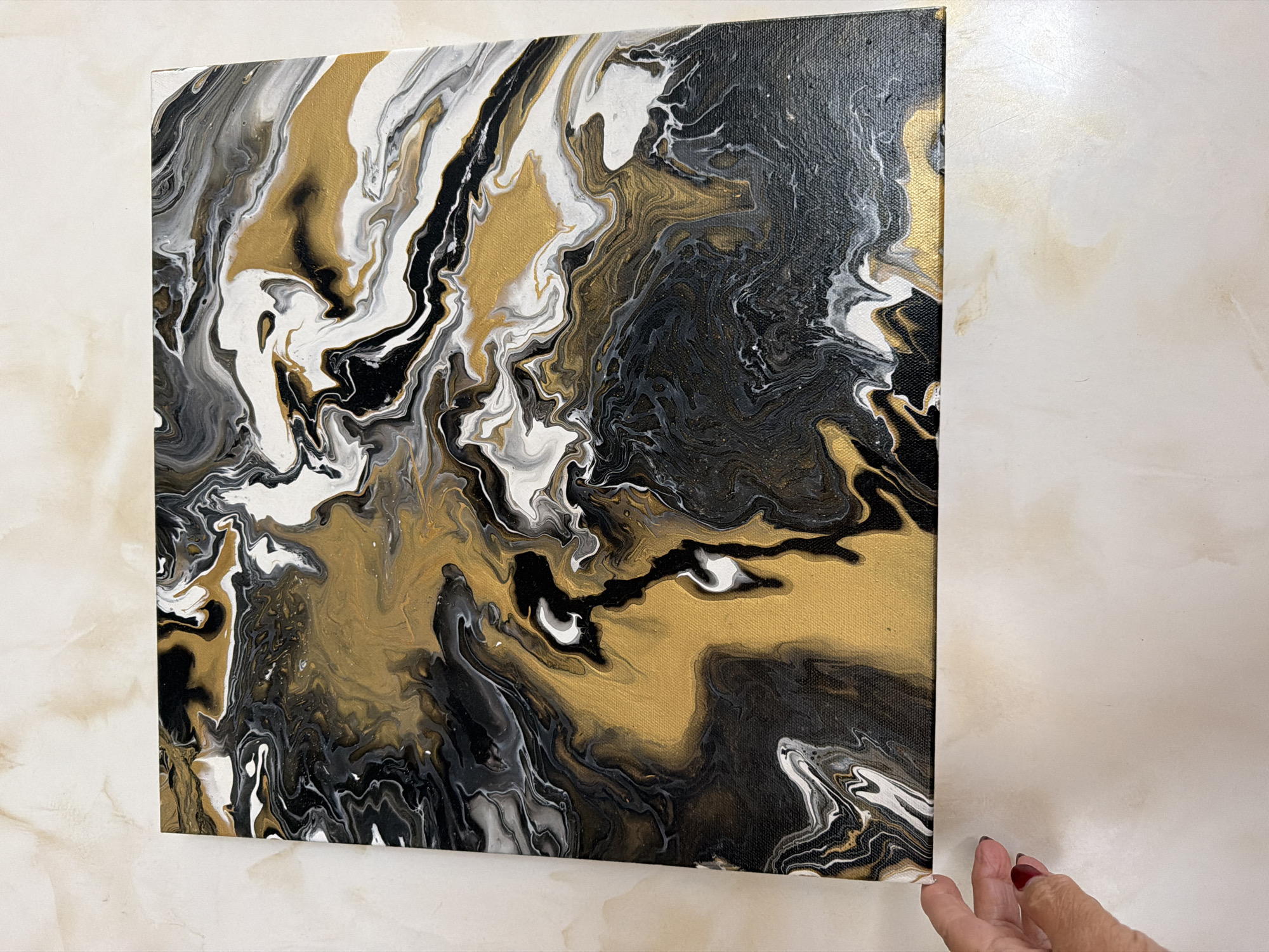 A close-up, high-angle shot shows a person's left hand with dark red nail polish lightly touching the bottom-left corner of a canvas. The canvas is a pour painting with abstract swirls of black, white, and metallic gold paint, resembling marble or flowing liquid. The painting is positioned on a light-colored, textured surface that appears to be a table or countertop. The background is out of focus, showing a subtle marbled pattern. The overall impression is of an art piece being presented or displayed.