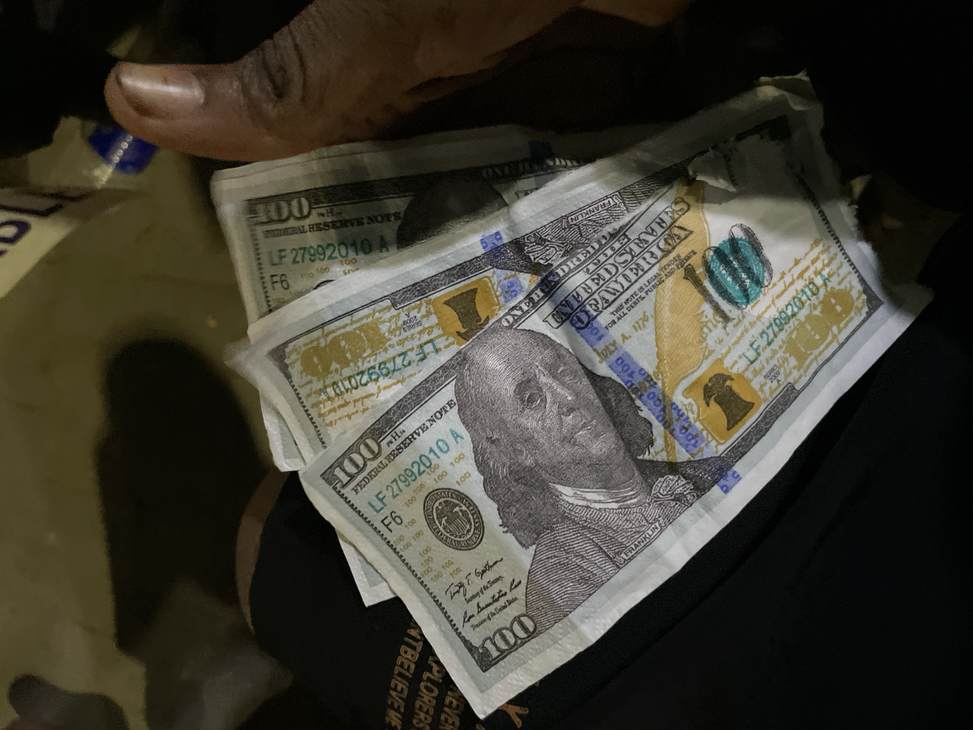 A hand is holding a fan of US hundred-dollar bills. The bills appear to be authentic, with visible security features. The background is dark and out of focus, suggesting an indoor setting or nighttime. There is a portion of black fabric visible at the bottom left, possibly clothing. The context provided about Lekki, Nigeria is not supported by any visual elements in the image itself.