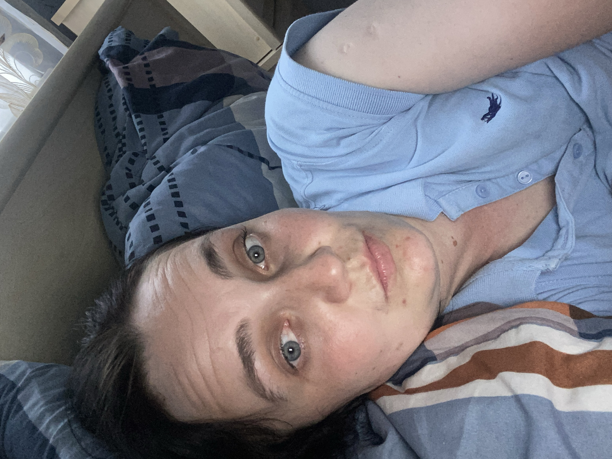 A low-angle, overhead shot shows a person lying on their back in bed. The person is facing the camera, and their head is tilted to their right. Their eyes are open and looking towards the camera. They have light blue eyes and their skin is fair, with some freckles. They are wearing a light blue polo shirt with a dark blue embroidered logo on the left chest, and the collar is visible. The person's hair is dark brown and appears to be shoulder-length.
The person is lying on a bed with patterned bedding. The blanket closest to the person has horizontal stripes in blue, white, brown, and gray. The blanket further away has a pattern of dark blue squares and rectangles on a lighter blue background.
To the left of the person, a wooden headboard or furniture is visible. A sheer curtain with a floral pattern can be seen through a window behind the headboard. The lighting suggests it is daytime, with soft, natural light illuminating the scene. The overall mood appears relaxed and intimate. No text is visible in the image.