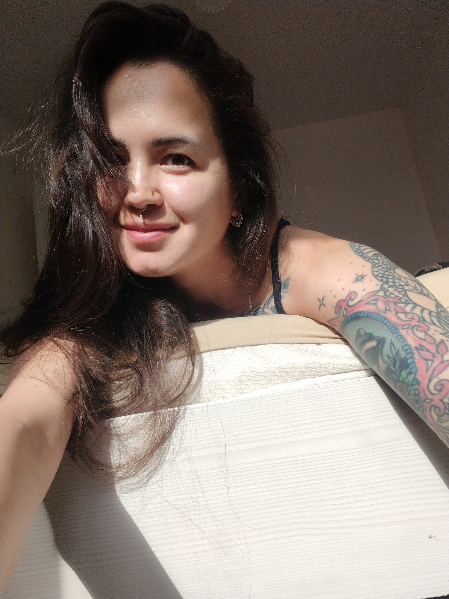 This media file is a close-up, self-portrait photograph of a young woman lying on a bed. The woman has long, dark, wavy hair that falls across her face and shoulders. She is wearing a dark camisole or tank top. Her left arm, which is visible, is heavily tattooed with colorful designs, including what appears to be a depiction of an animal and intricate patterns. She has a nose ring with a small charm and is wearing small hoop earrings.
The woman is looking directly at the camera with a slight smile. Her expression appears relaxed and content. The lighting suggests it is daytime, with sunlight casting a strong shadow across the bed and the woman's arm, indicating the sun is positioned to the left of the frame. The background is a plain, neutral-toned wall, with a hint of the room's interior. The bed appears to be covered with a textured, light-colored fabric, possibly a sheet or duvet.
The overall mood of the image is intimate and casual, capturing a candid moment. There is no visible text in the image. Given the location context provided as "Noviy, Russia," this scene is likely taking place indoors in a residential setting in Russia.
