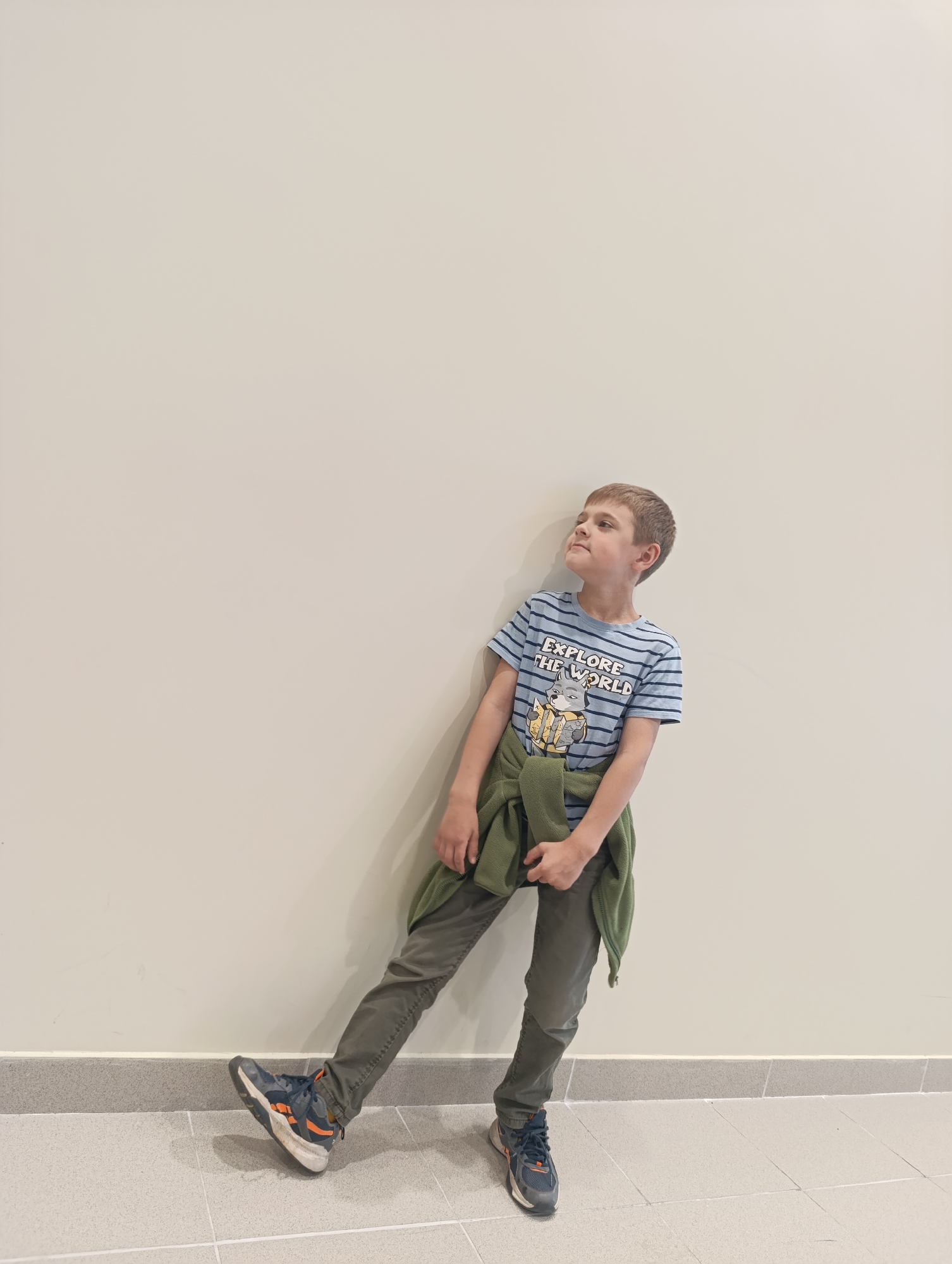A young boy stands leaning against a plain, cream-colored wall. He is wearing a blue and white striped t-shirt with a graphic print of a cartoon character and the text "EXPLORE THE WORLD." A green fleece jacket is tied around his waist. He has on dark grey pants and blue and orange athletic shoes. His left leg is extended forward, and his right leg is bent. The boy is looking upwards and to his right. The floor is tiled in a light grey color. The scene appears to be indoors, possibly in a public building, with a clean and minimalist environment. There is no indication of the city of Domodedovo, Russia.