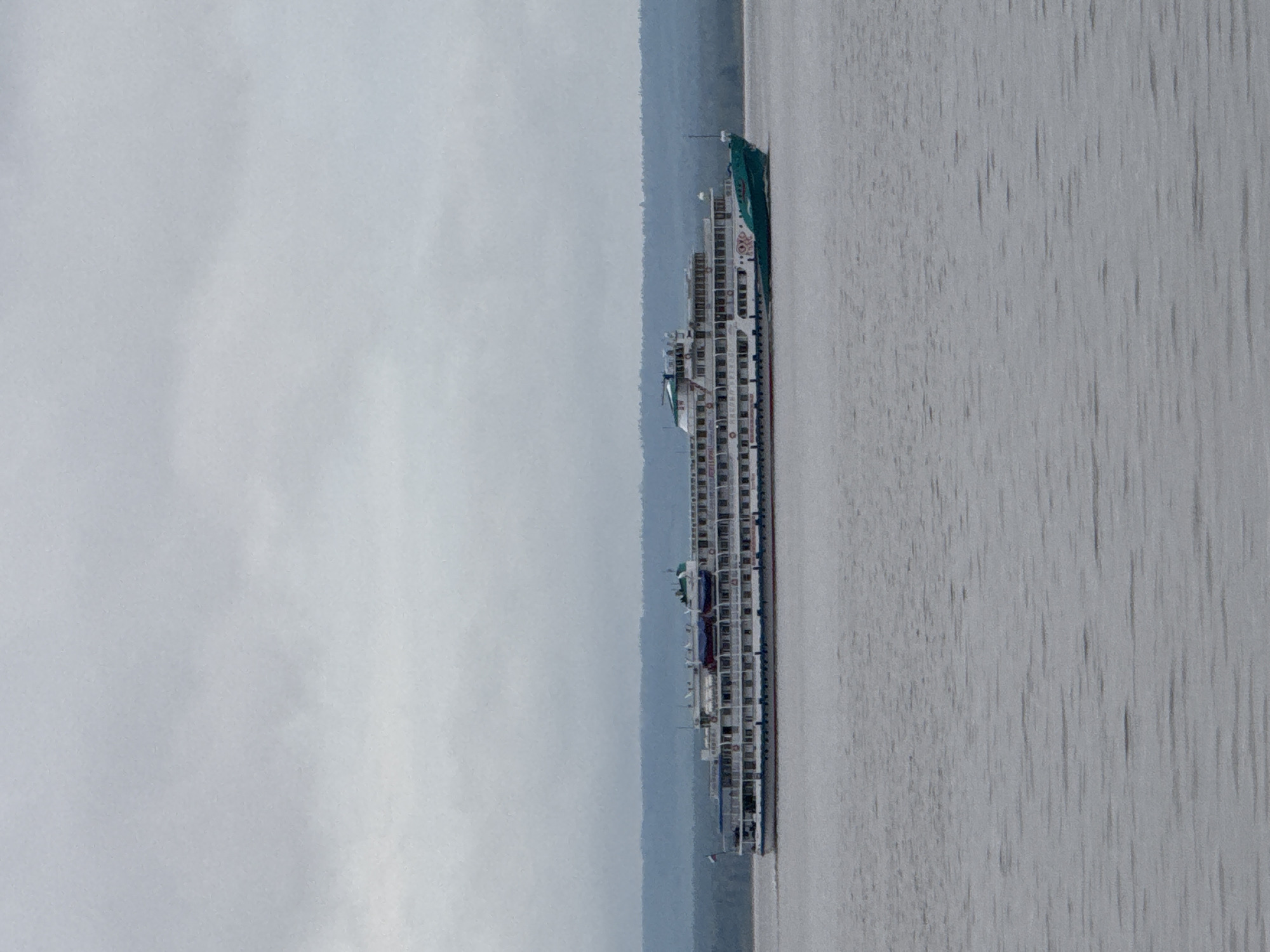 The image depicts a multi-deck cruise ship docked in the city of Chastyye, Russia. The ship is white with multiple rows of rectangular windows. The background includes a hazy gray sky and water. Some shoreline or land is visible on the left side.